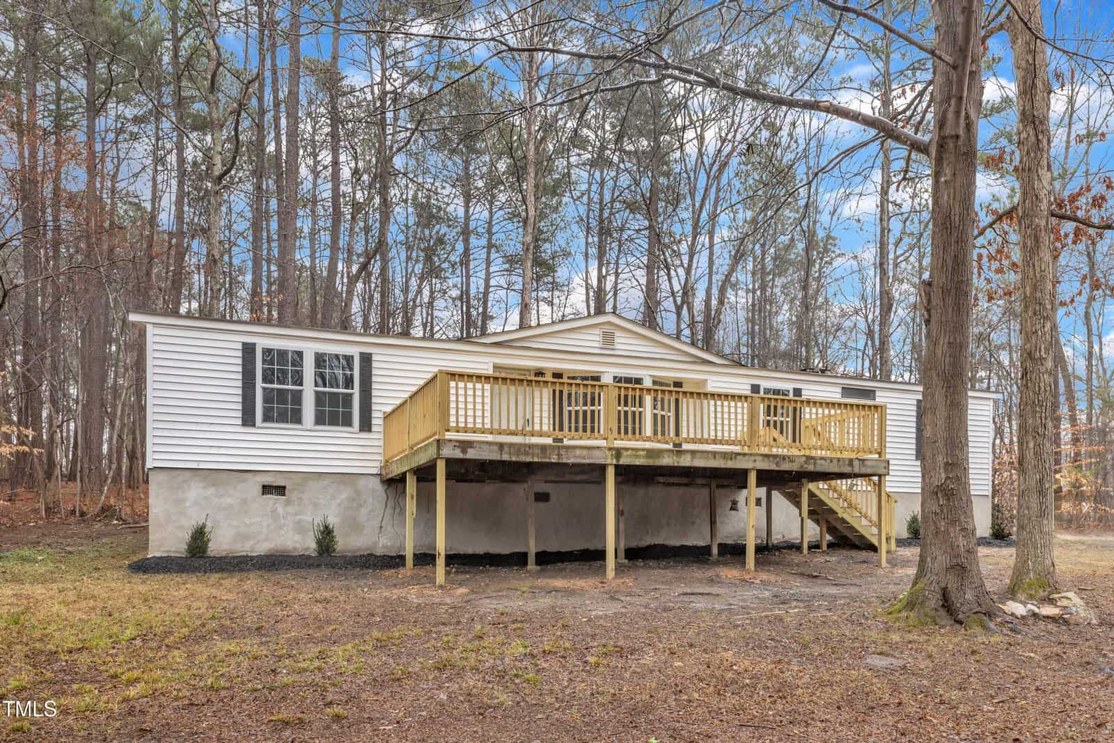 A white manufactured home on 3519 Topaz Street features a wooden deck with railing and steps, nestled in a wooded area. Bare-branched trees encircle the property, all under a partly cloudy sky typical of charming Wake Forest.