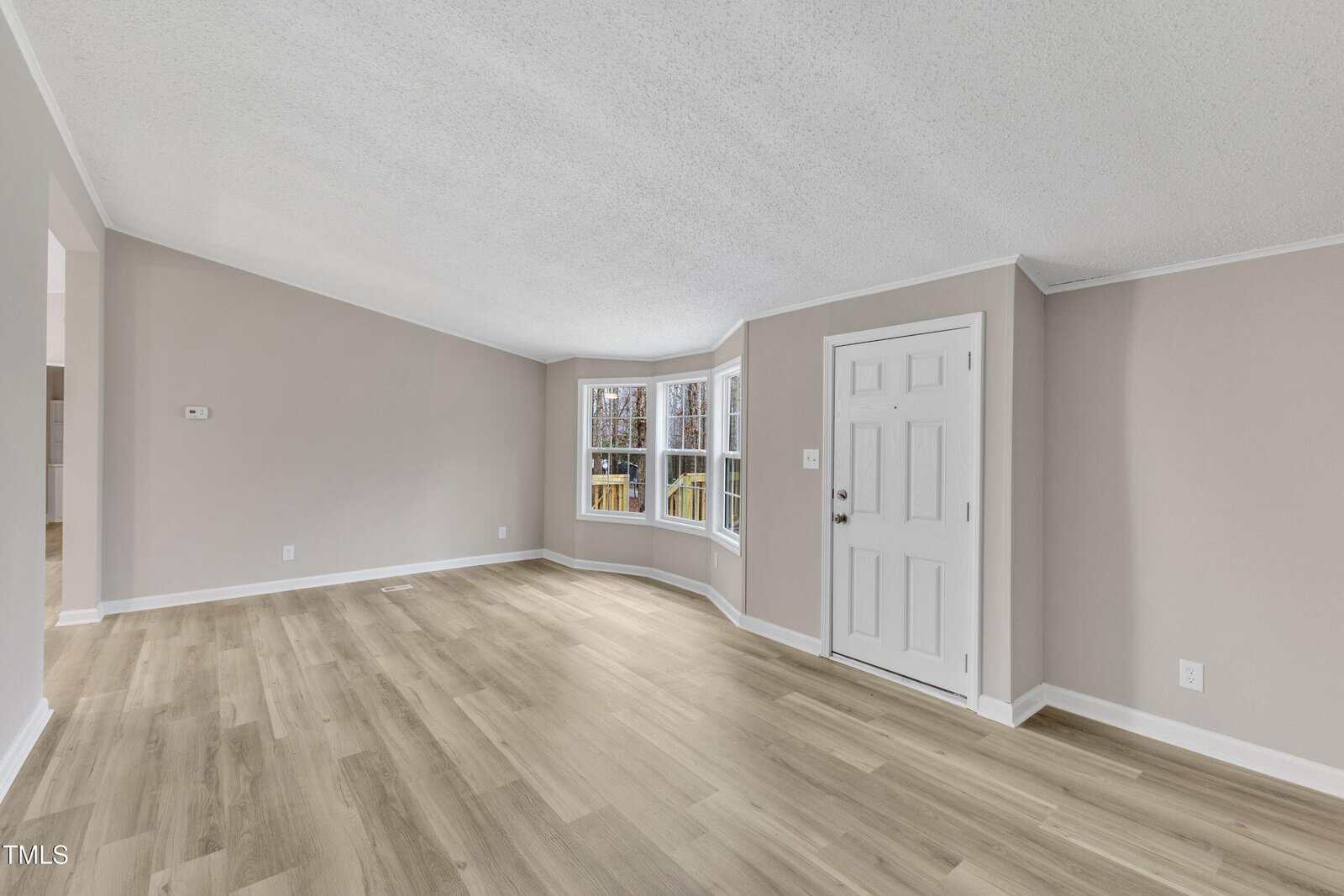 A spacious living room with light wood flooring, gray walls, and a white ceiling. The room features a bay window on the right with a view of trees and a white door near the window, leading outside. The space is empty and well-lit.