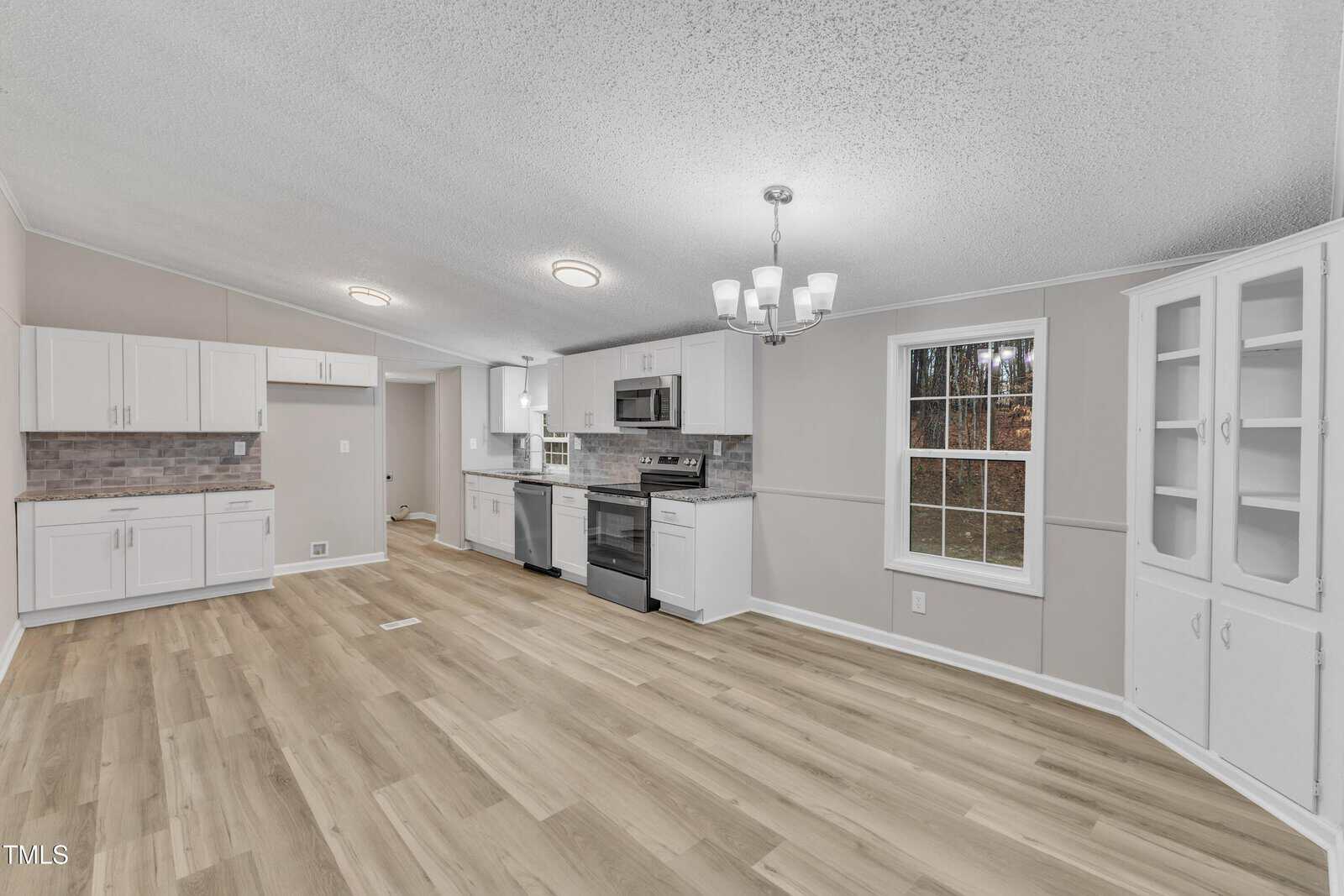 A modern kitchen and dining area with white cabinets, stainless steel appliances, and a light wood floor. There's a window on the right, letting in natural light, and a chandelier hanging above. A small built-in corner cabinet is visible on the right wall.