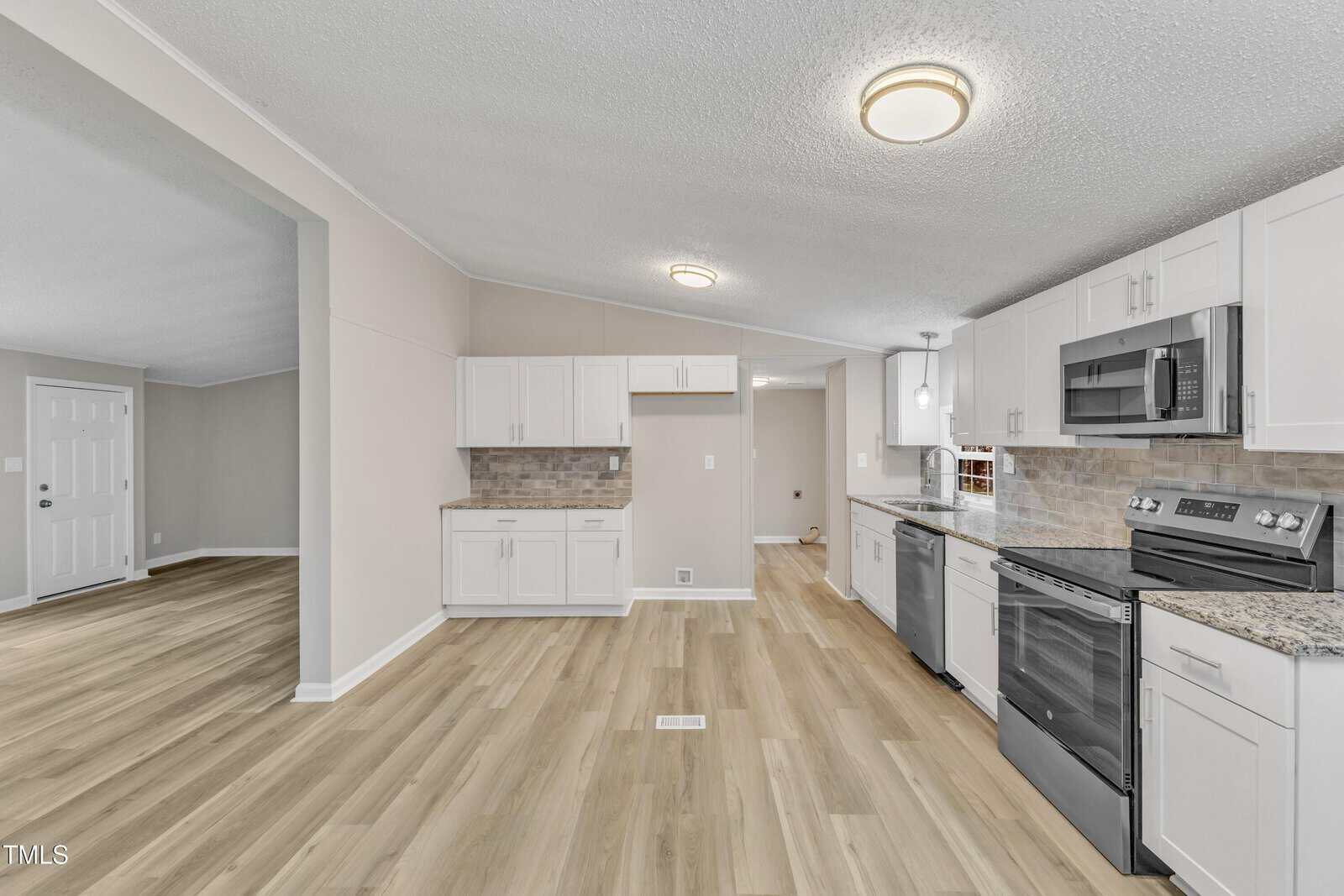 A modern kitchen with white cabinets, stainless steel appliances, and a gray tile backsplash. The room has light wood flooring and a ceiling light fixture. An open doorway leads to an adjacent room with matching flooring and a closed white door.