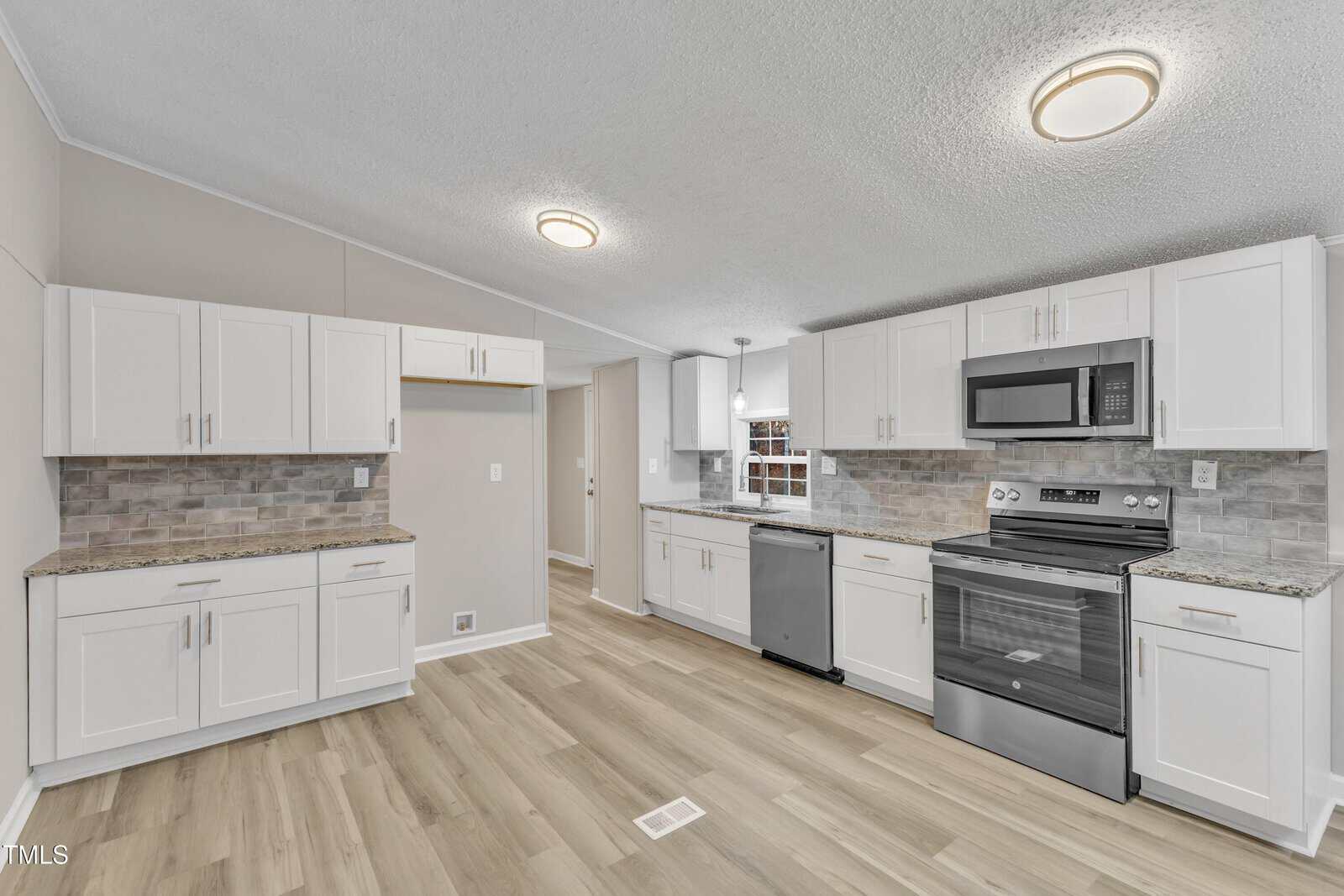 A modern kitchen with light wood flooring and white cabinets. It features a stainless steel stove, microwave, and dishwasher. The countertops are granite with a gray tile backsplash. Ceiling lights illuminate the space.