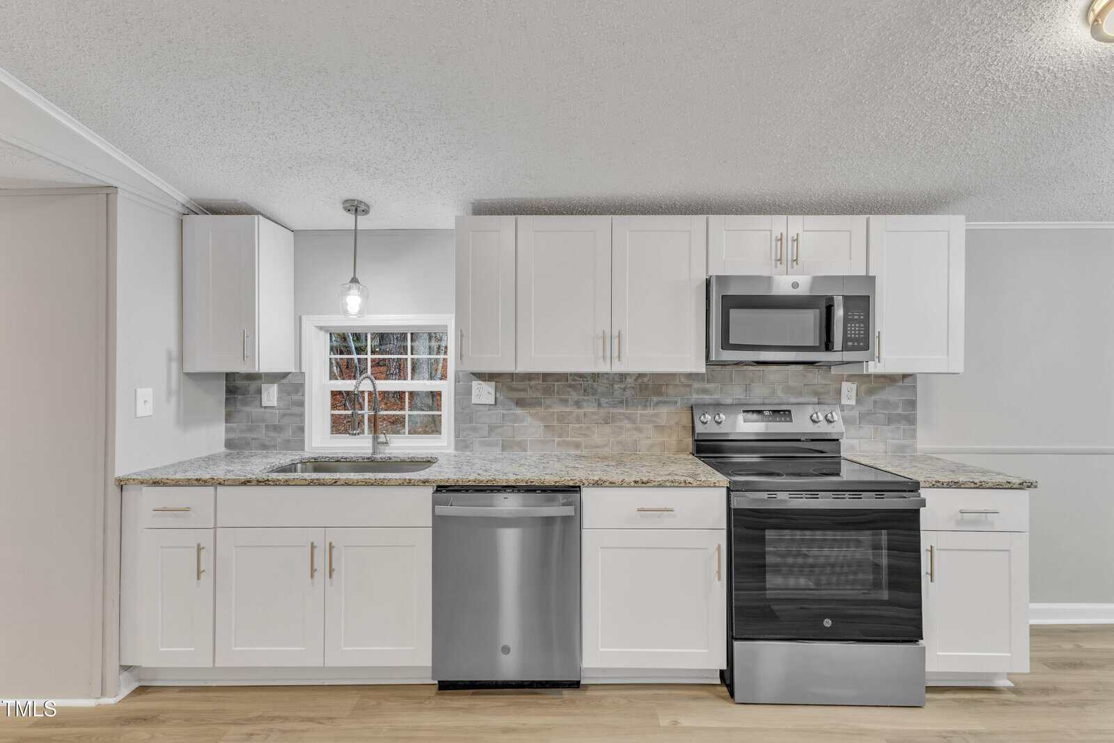 A modern kitchen with white cabinets, stainless steel appliances, and a tile backsplash. There is a window above a double sink, a microwave above the stove, and a dishwasher next to it. The floor appears to be wooden.