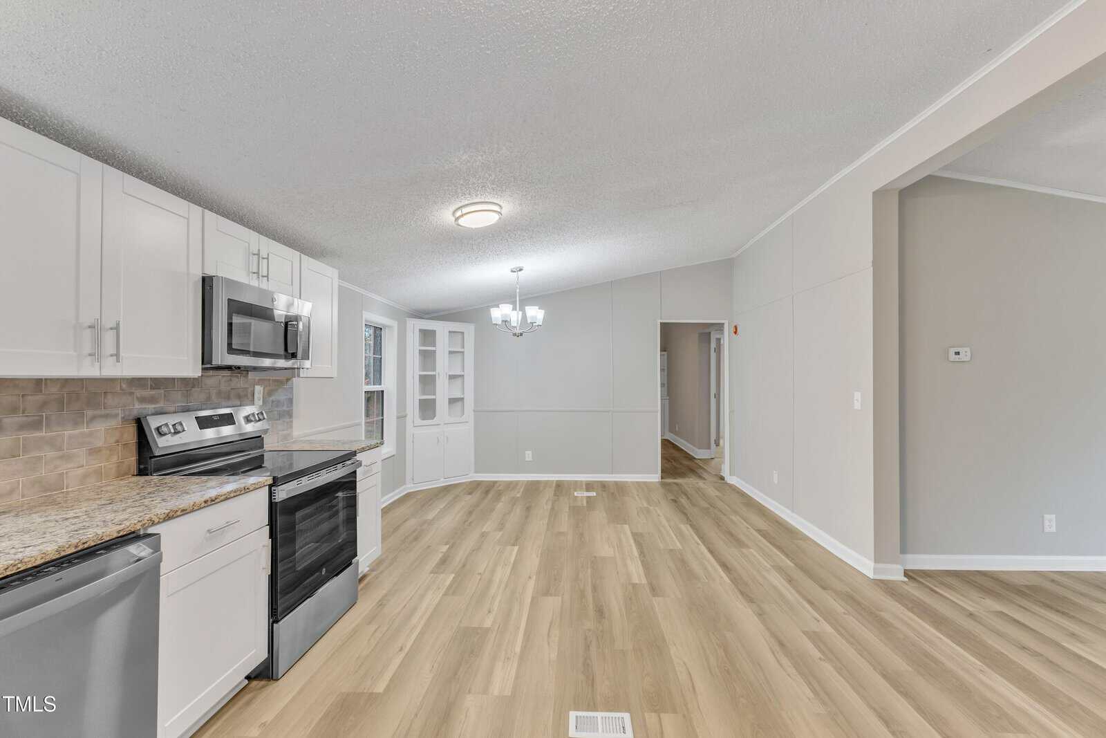 A modern kitchen and dining area with wooden flooring. The kitchen features white cabinets, stainless steel appliances, and a brick-patterned backsplash. There's a ceiling light fixture, and an open dining space with a small built-in cabinet.