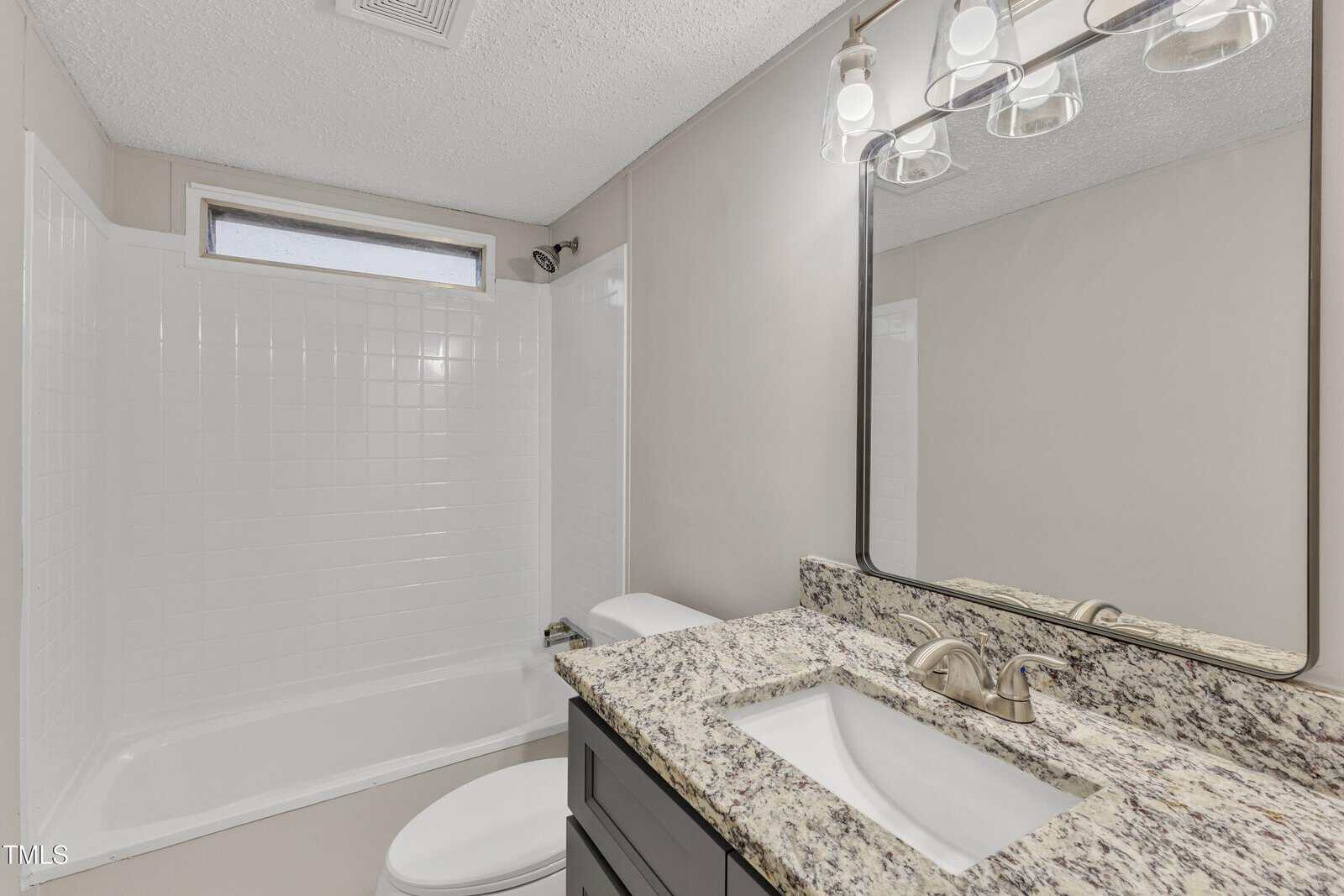 Bathroom with a granite countertop and a sink. Above the sink is a large mirror with three light fixtures. To the left is a white bathtub with tiled walls and a small window. The walls are light-colored.