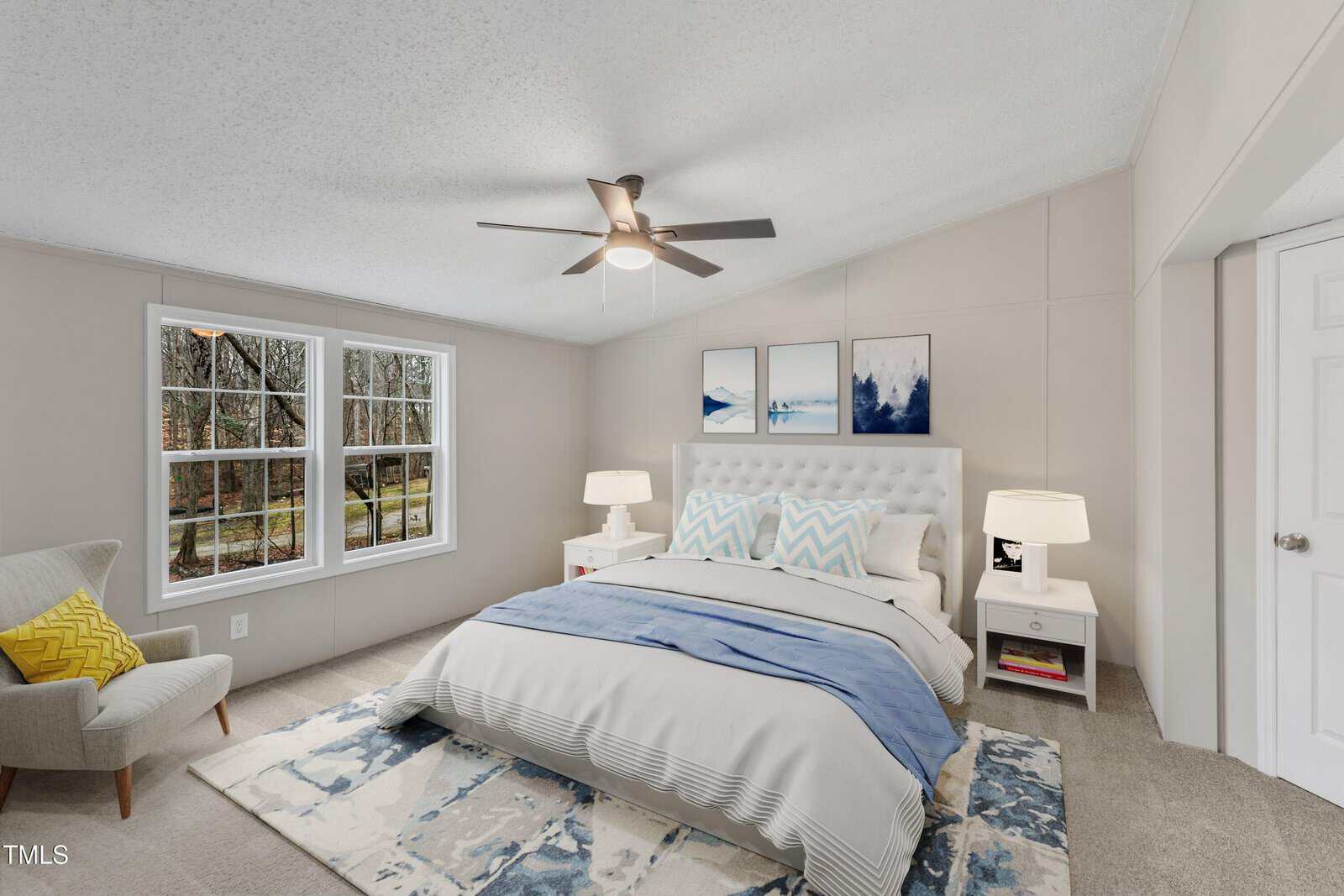 A bedroom featuring a bed with white and blue bedding, two nightstands with lamps, a ceiling fan, a patterned rug, and a gray armchair. A large window offers a view of trees, and there are three abstract paintings on the wall.