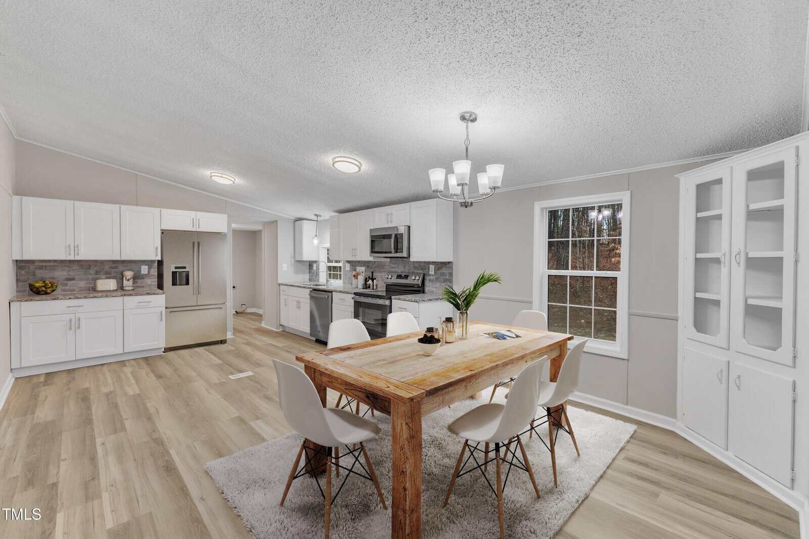 A modern kitchen and dining area with hardwood floors. Features a wooden dining table with six white chairs on a gray rug. The kitchen has white cabinets, stainless steel appliances, and a window with a view of trees. A chandelier hangs above the table.
