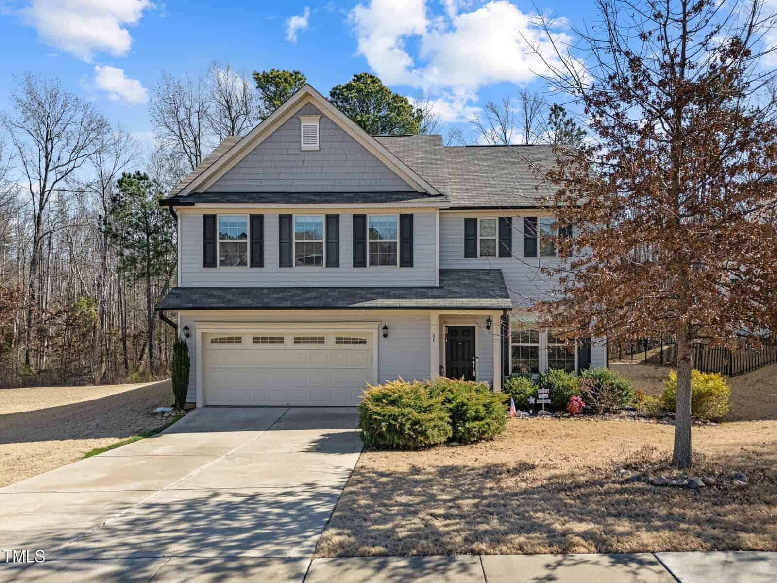 Two-story suburban house with gray siding, black shutters, and a two-car garage. The front yard has dry grass, some shrubs, and a leafless tree. The sky is partly cloudy, and trees line the background.