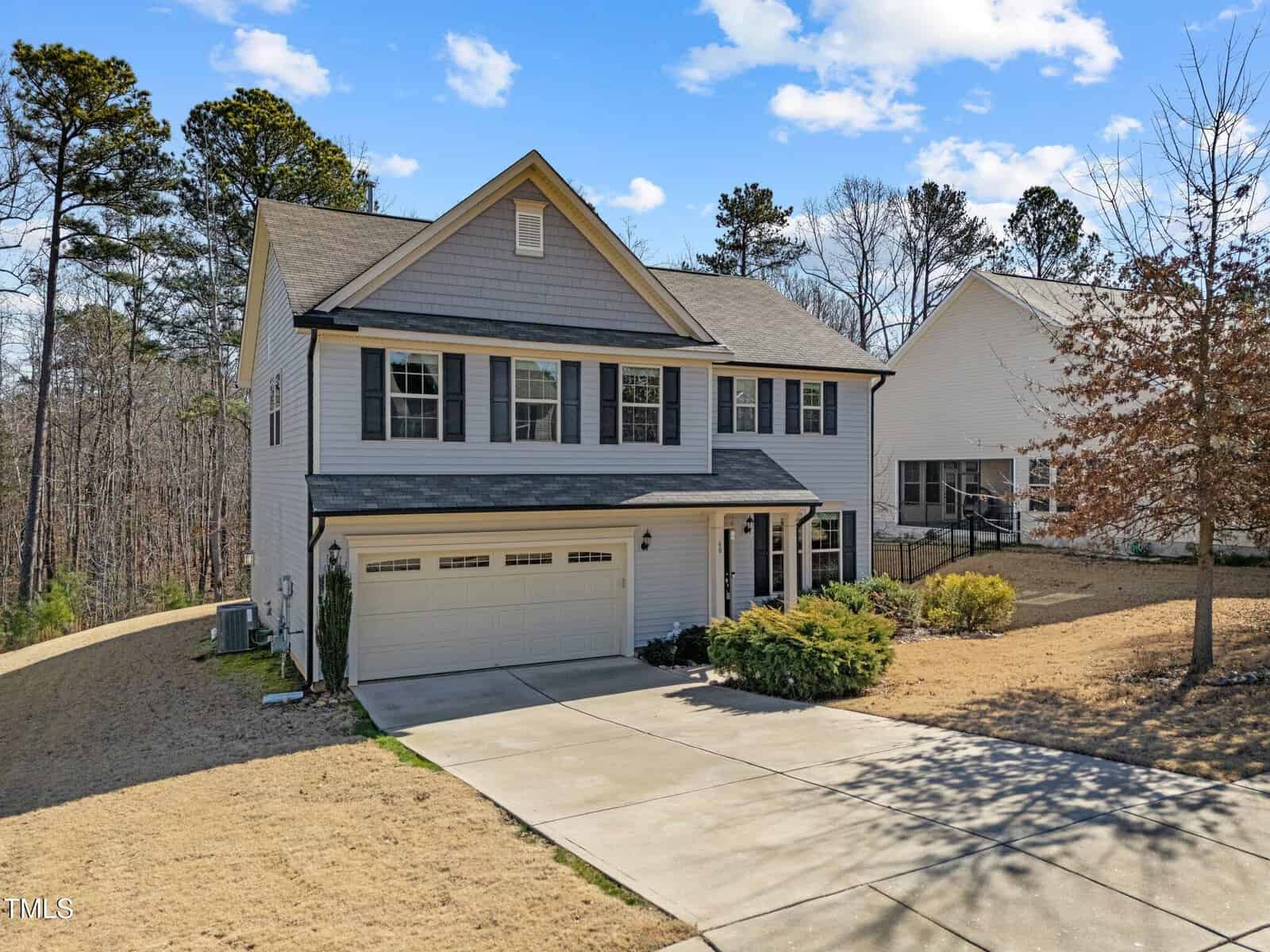 Two-story suburban house with gray siding, black shutters, and a two-car garage. A concrete driveway leads to the garage, and there is minimal landscaping with some bushes and a sloped, patchy lawn. Trees are in the background.