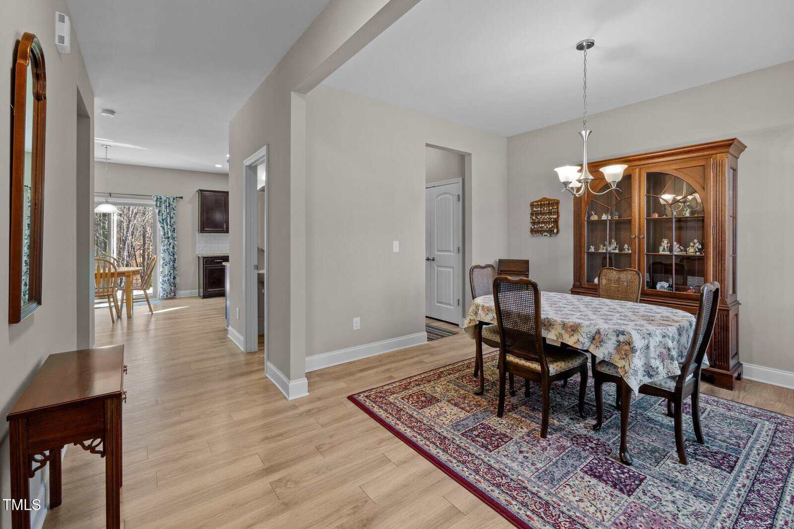 A dining room with a wooden table and six chairs, a floral tablecloth, a china cabinet, and a patterned rug. Adjacent hallway leads to a kitchen and a breakfast area with a table and chairs near glass doors.
