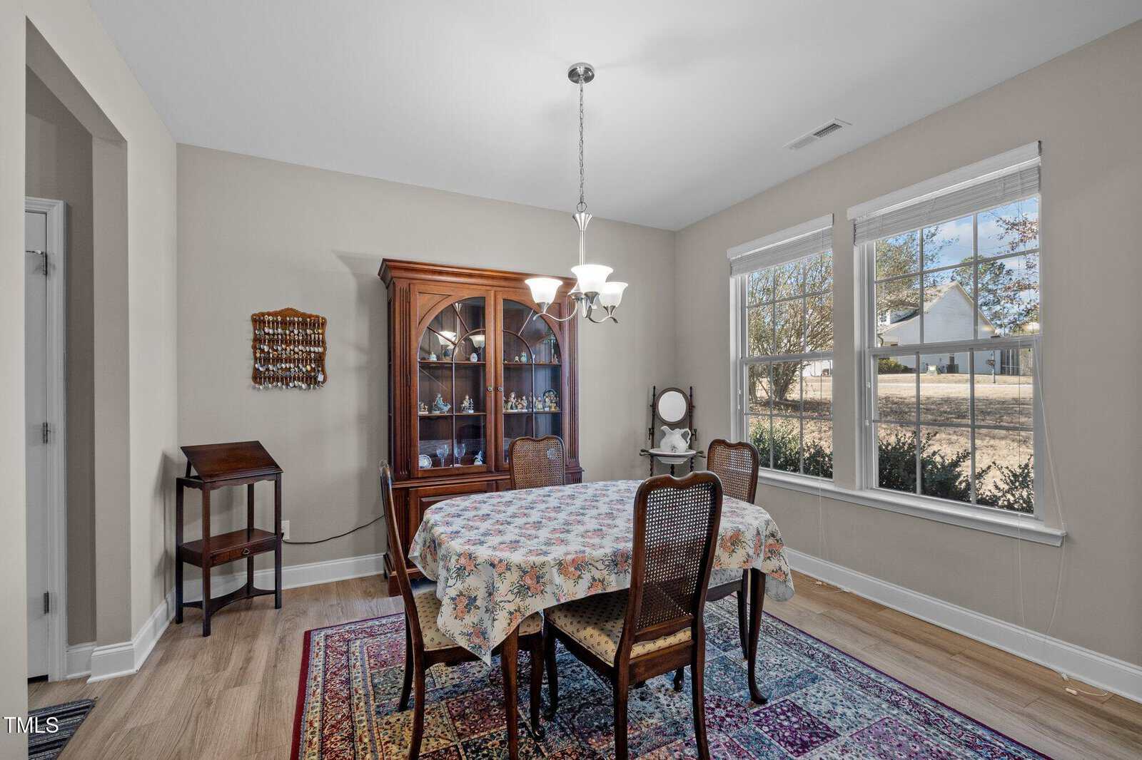 A dining room with a floral tablecloth on a wooden table, four chairs, a rug, a china cabinet, a small shelf, and large windows letting in natural light. Walls are beige and the floor is light wood.