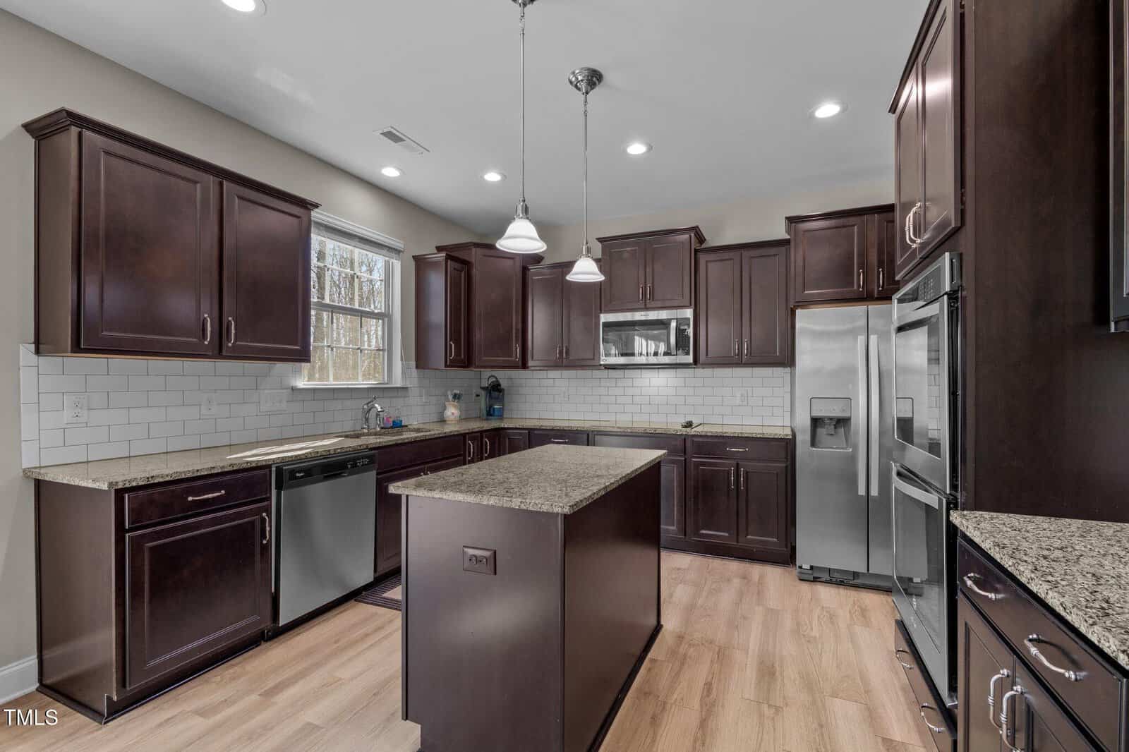 Modern kitchen with dark wood cabinets, stainless steel appliances, a center island, granite countertops, white subway tile backsplash, light wood flooring, and two pendant lights. A window above the sink lets in natural light.