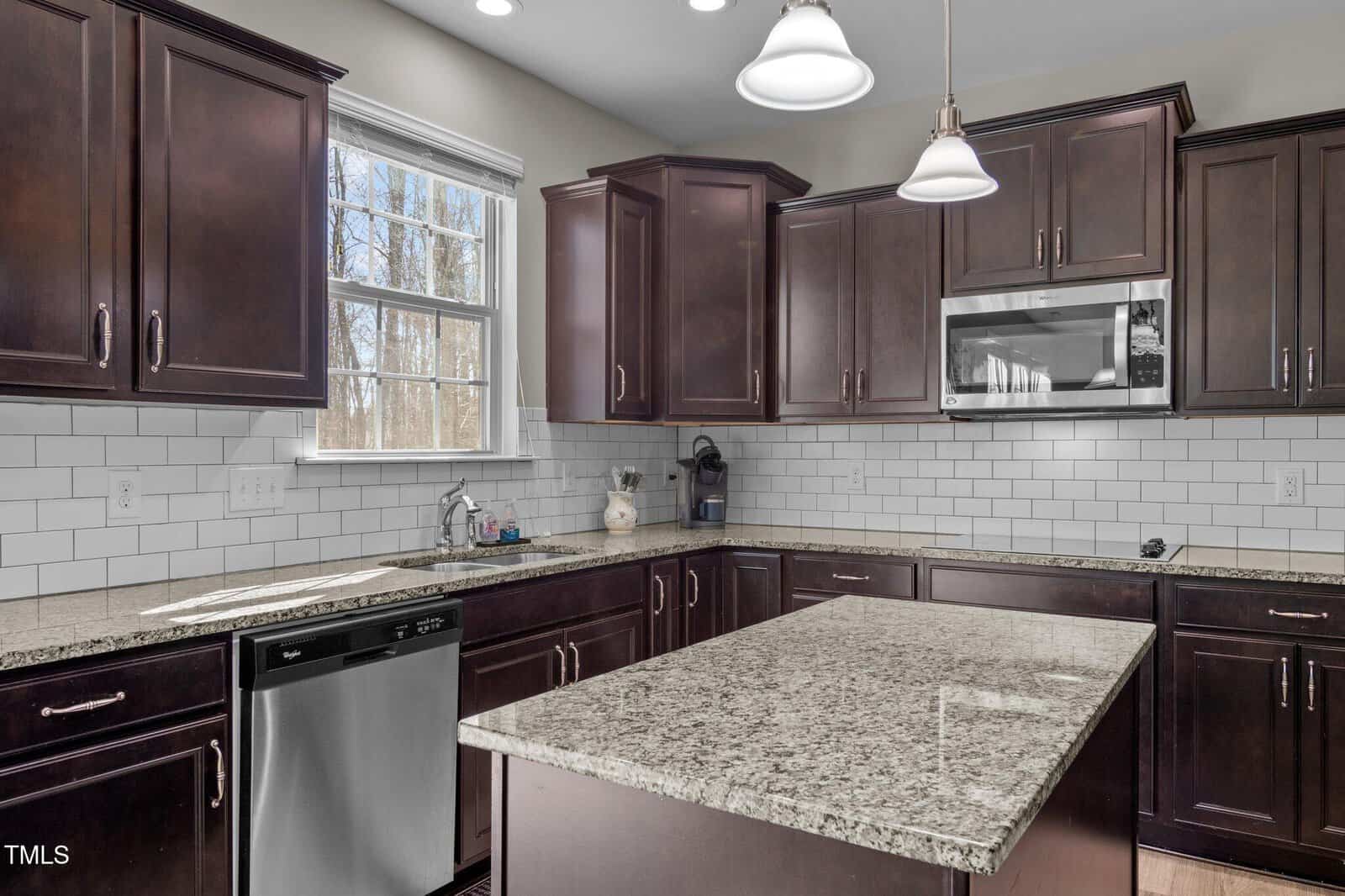 Modern kitchen with dark wood cabinets, white subway tile backsplash, granite countertops, stainless steel dishwasher, microwave, double pendant lights, and a window above the sink letting in natural light.