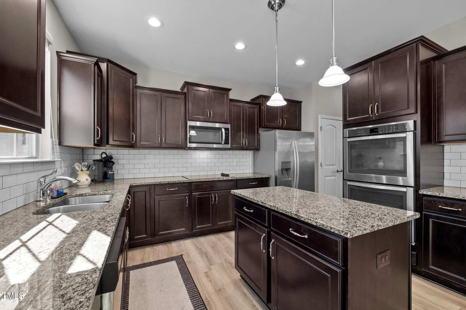 Modern kitchen with dark wood cabinets, stainless steel appliances, granite countertops, a kitchen island, white subway tile backsplash, three pendant lights, and wood flooring. Sunlight streams in through a window above the sink.