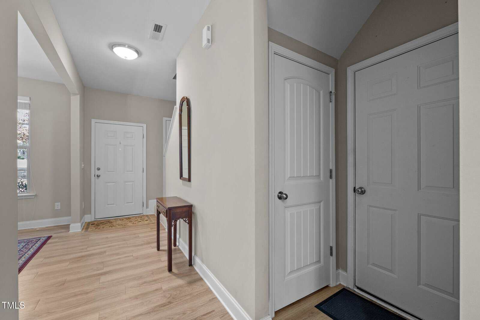 A hallway with light wood flooring, beige walls, a white front door, a white interior door, a small wooden table with a mirror above it, and a staircase partially visible in the background.