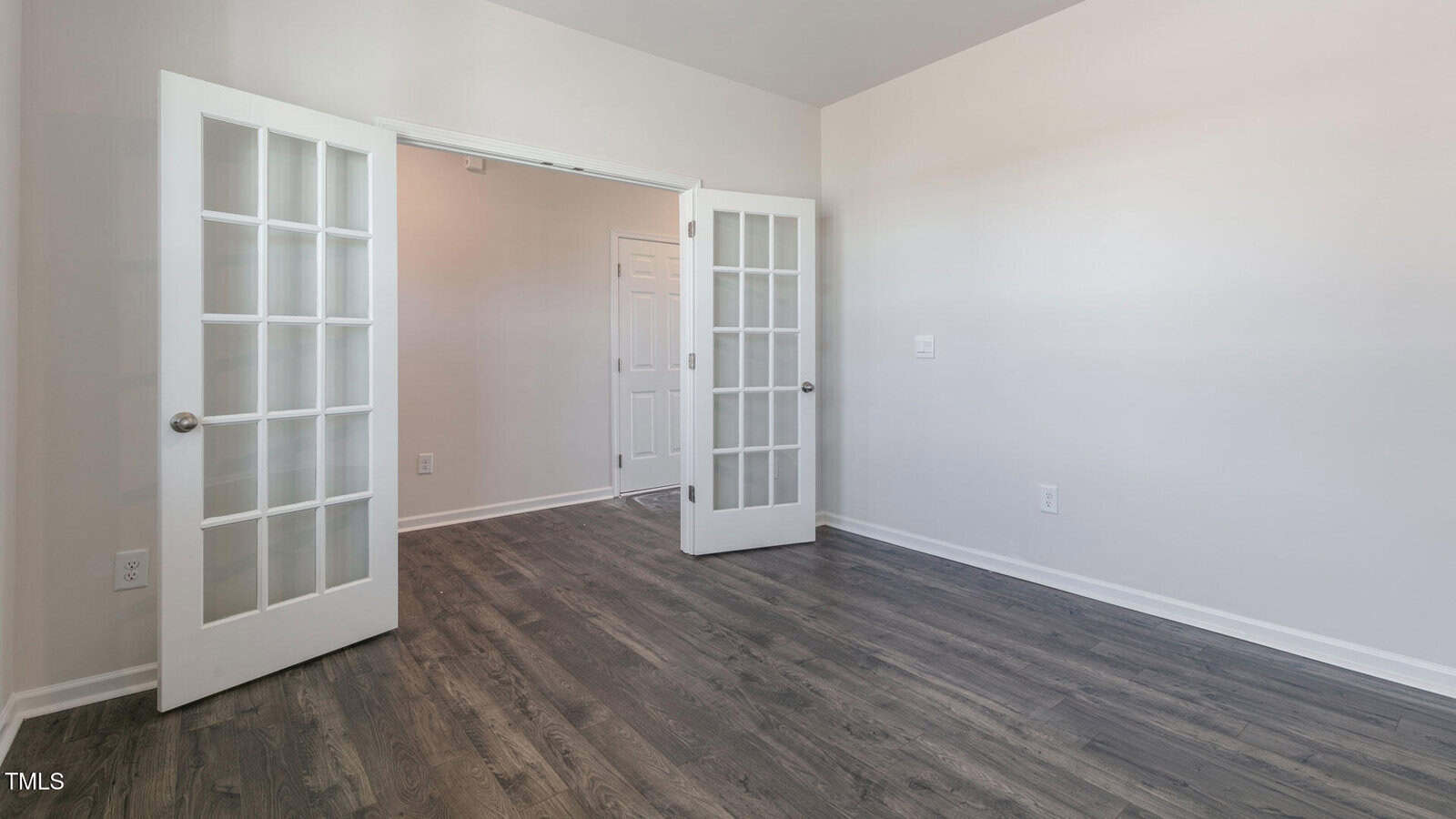 A room with light-colored walls, dark wood flooring, and double glass French doors opening to a hallway with a white door in the background. The room is empty and well-lit.
