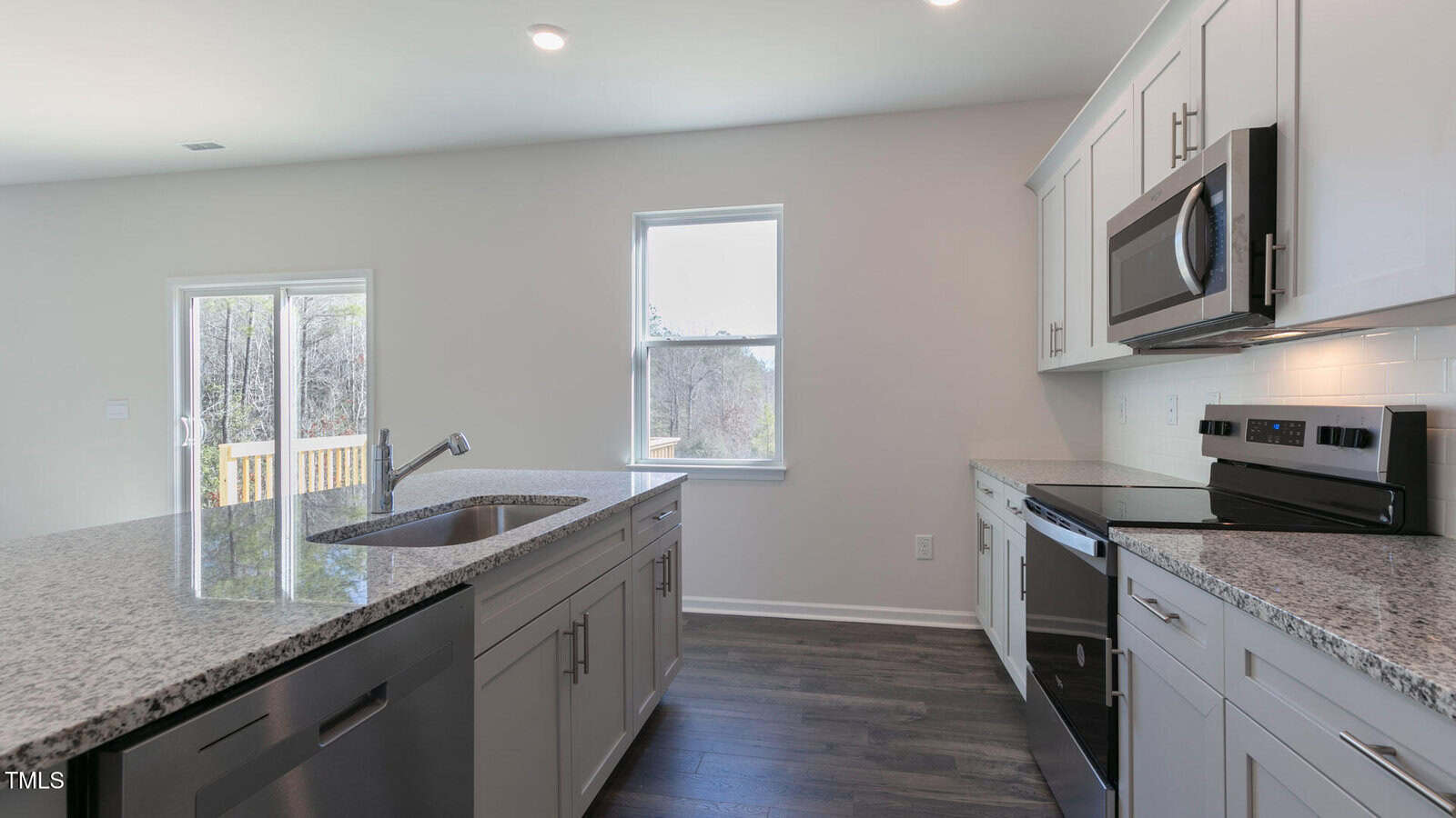 Modern kitchen with white cabinets, stainless steel appliances, an island with a sink, granite countertops, and hardwood floors. A window and glass door provide natural light and an outdoor view.