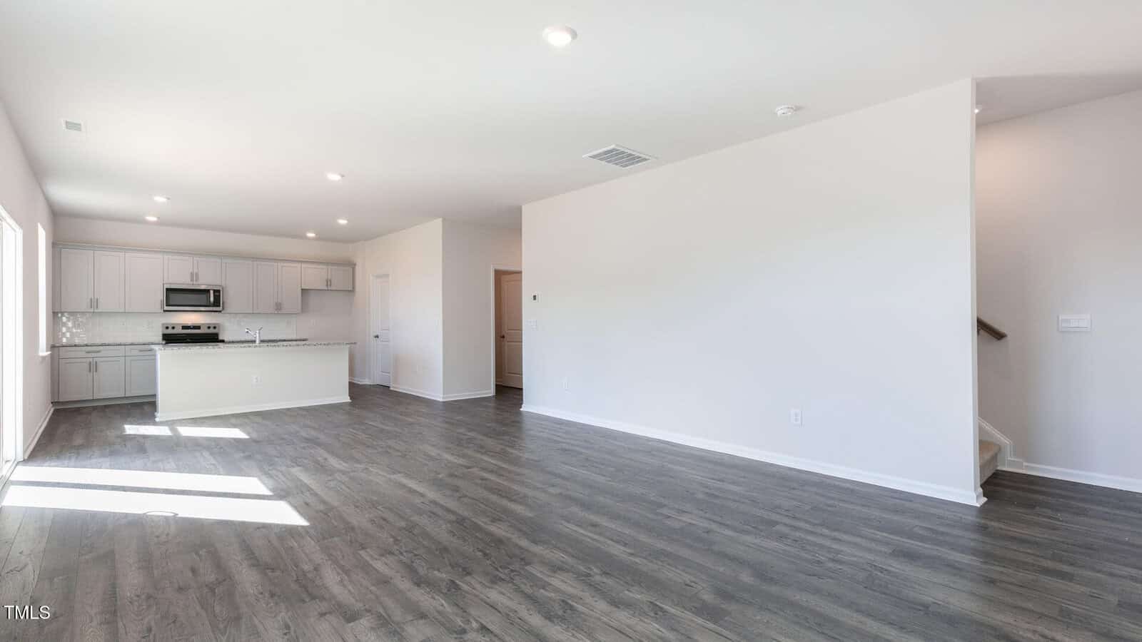 Open-plan room with light gray walls, wood-look flooring, and recessed lighting. Kitchen area has white cabinets, stainless steel appliances, and an island. Stairs are visible at the right side of the image.