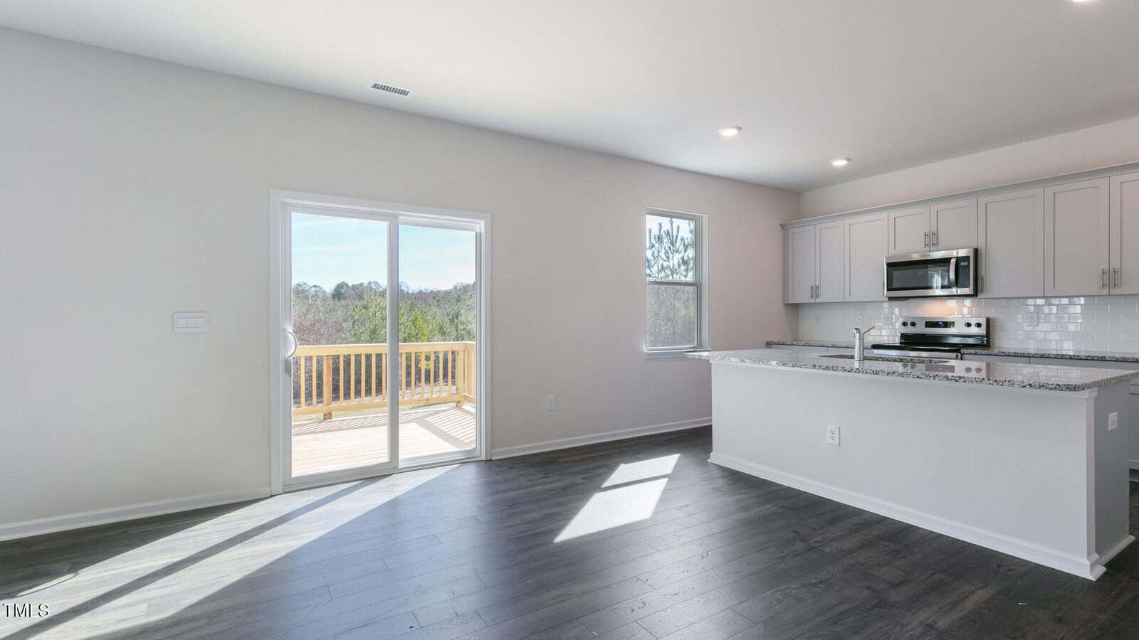 A modern kitchen with white cabinets, stainless steel appliances, granite countertops, and a breakfast bar. The room has dark wood flooring, a window, and sliding glass doors leading to a deck with a wooded view.