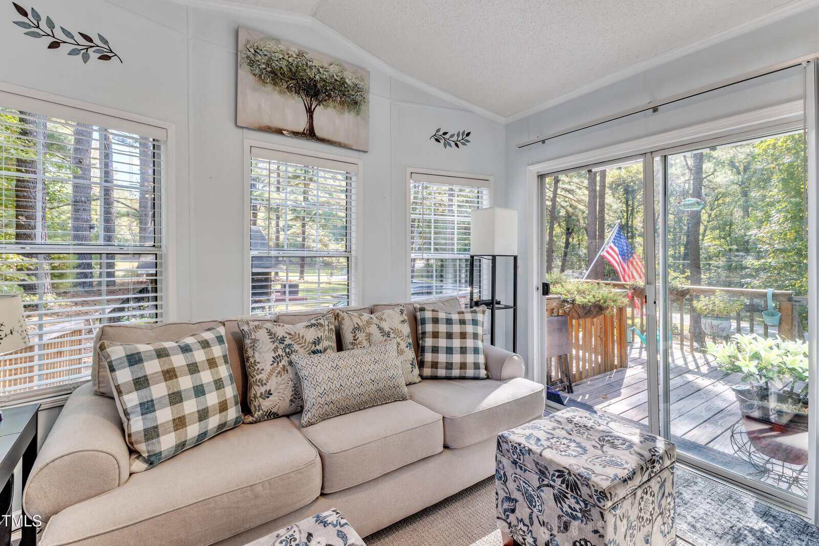 A beige sofa with patterned pillows is next to large windows and a glass door leading to a deck. The deck has plants and an American flag. Art and decor are on the walls, and sunlight streams into the bright room.