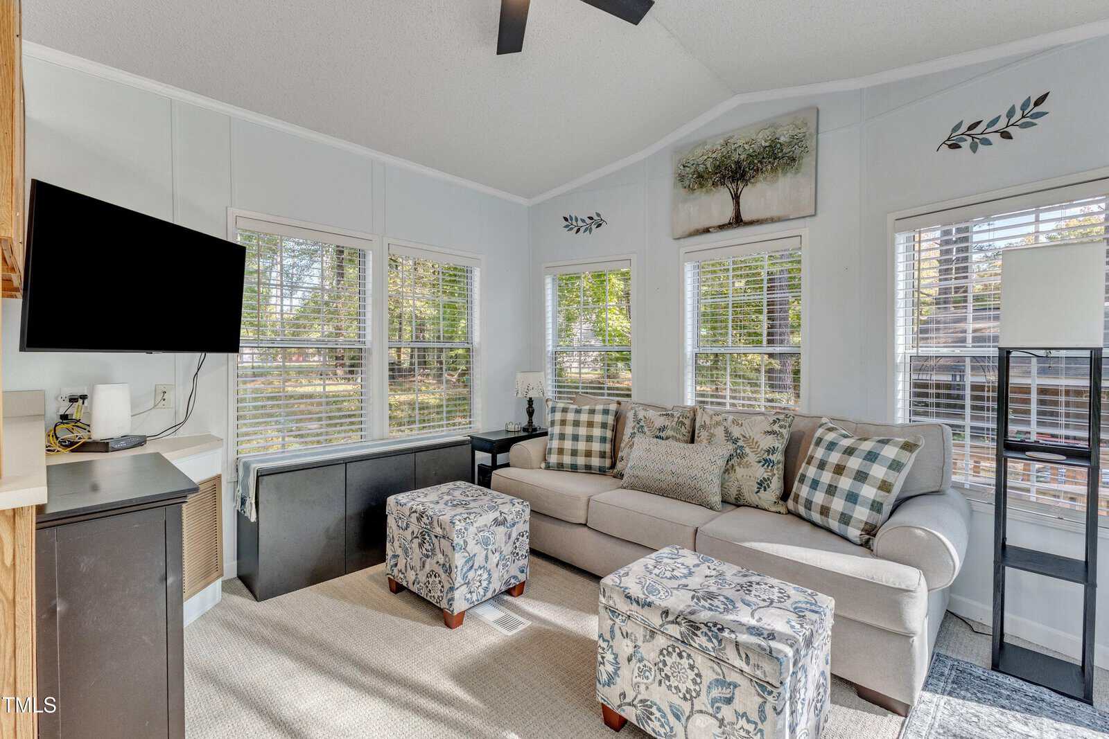 Bright sunroom with a beige sofa, patterned ottomans, wall-mounted TV, multiple windows, and light walls. A small desk sits by the window, and a wall art piece with a tree hangs above the sofa.