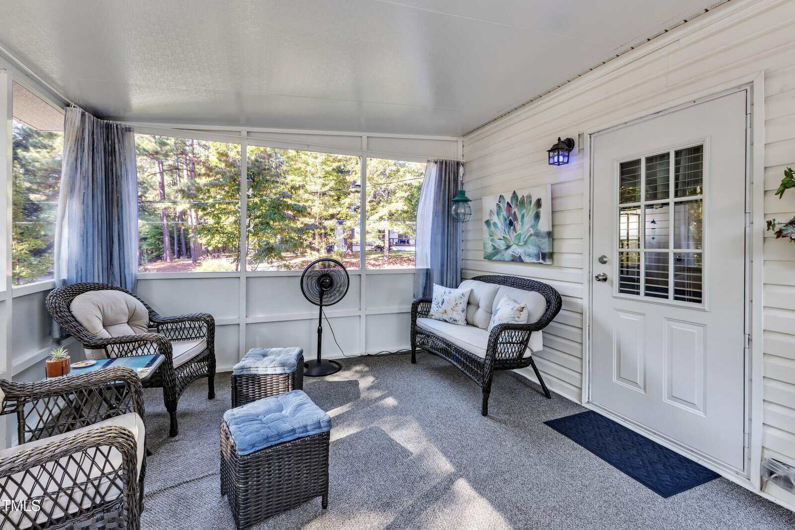 A screened-in porch with wicker furniture, cushioned chairs, a loveseat, small tables, a standing fan, a wall-mounted light, a decorative wall piece, and blue curtains, overlooking trees outside.