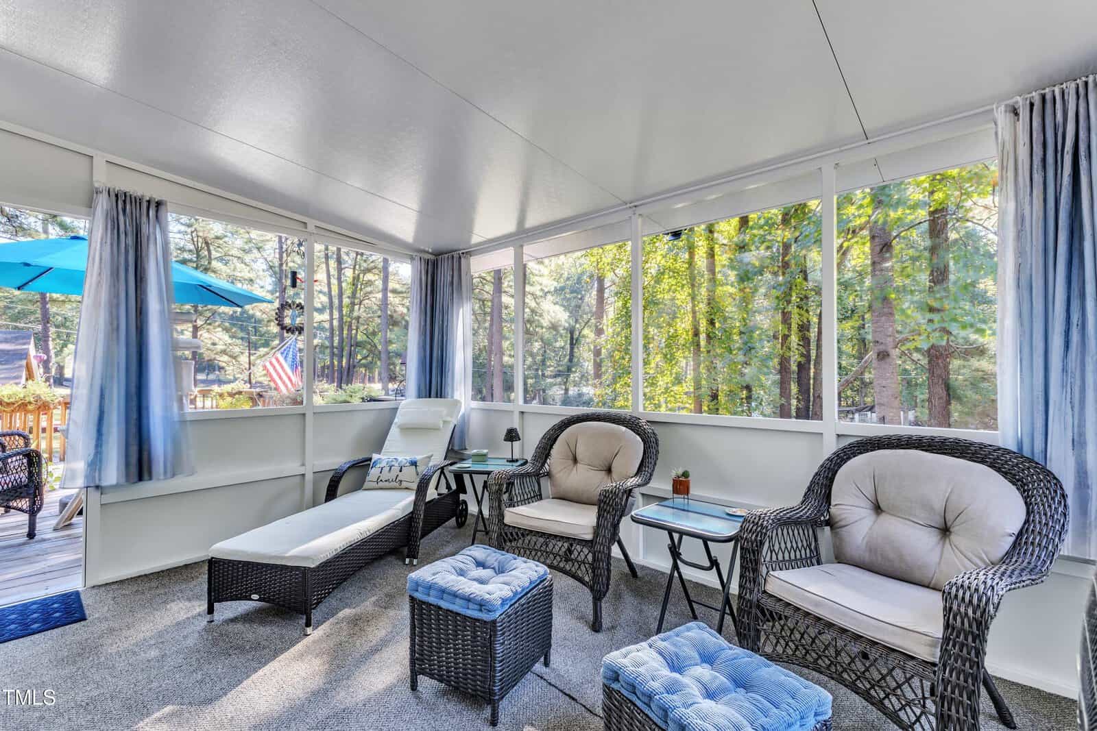 A screened-in porch with wicker chairs, a chaise lounge, small tables, cushioned ottomans, blue curtains, and large windows showing trees and an outdoor patio with an umbrella and American flag.