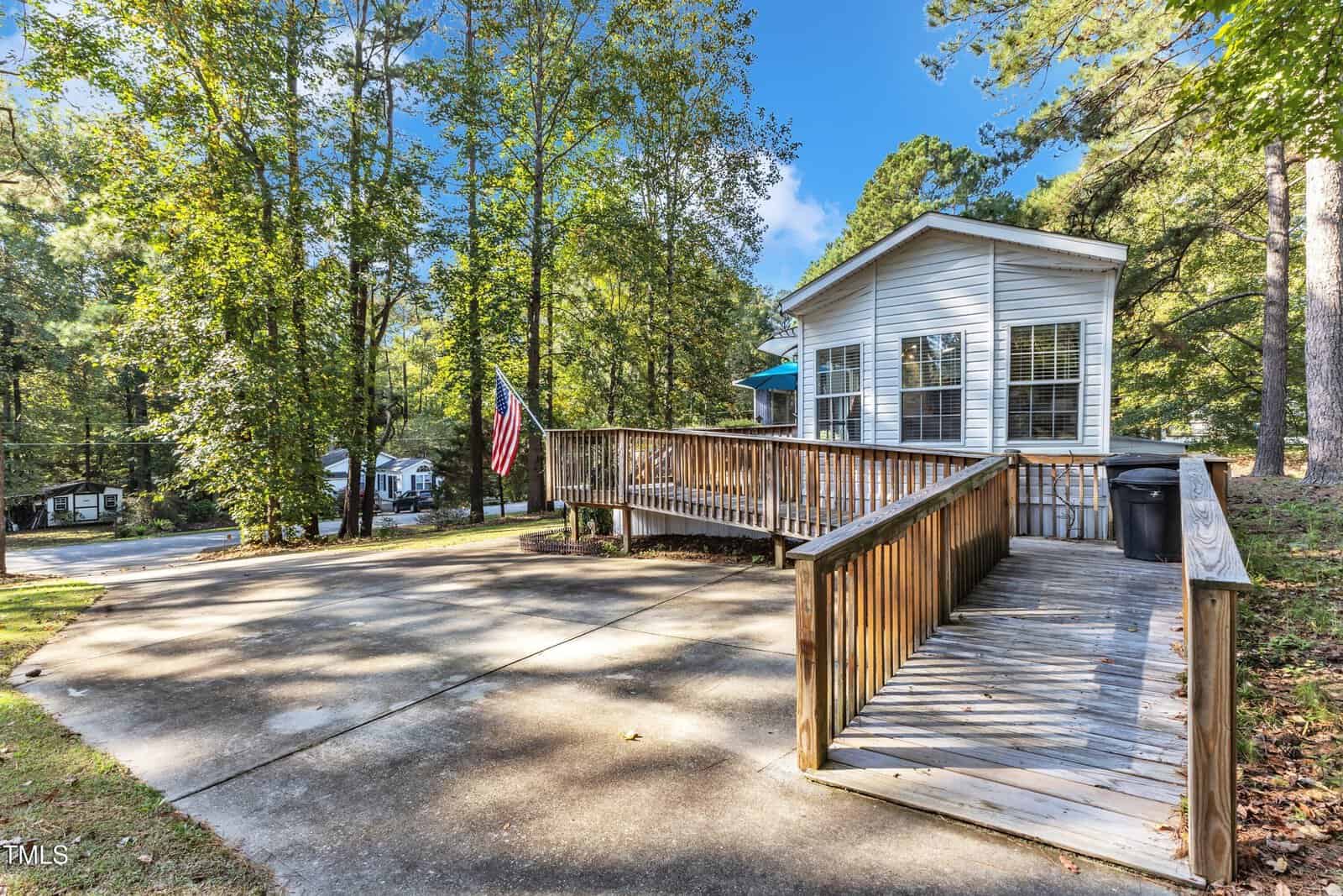 A white manufactured home with large windows sits among tall trees. A wooden ramp leads up to the entrance, and an American flag is displayed by the ramp. The foreground shows a wide concrete driveway.