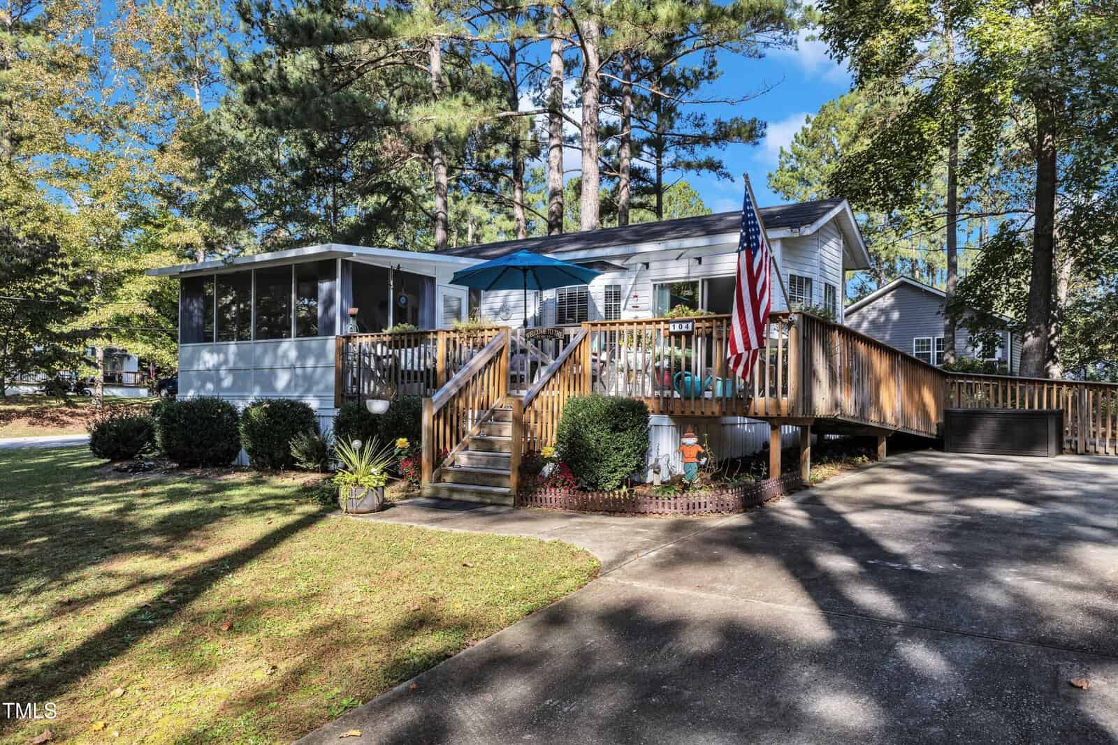 A single-story house with a large wooden ramp and deck, an American flag, patio furniture, screened porch, and surrounded by trees and shrubs on a sunny day. A curved driveway leads to the entrance.