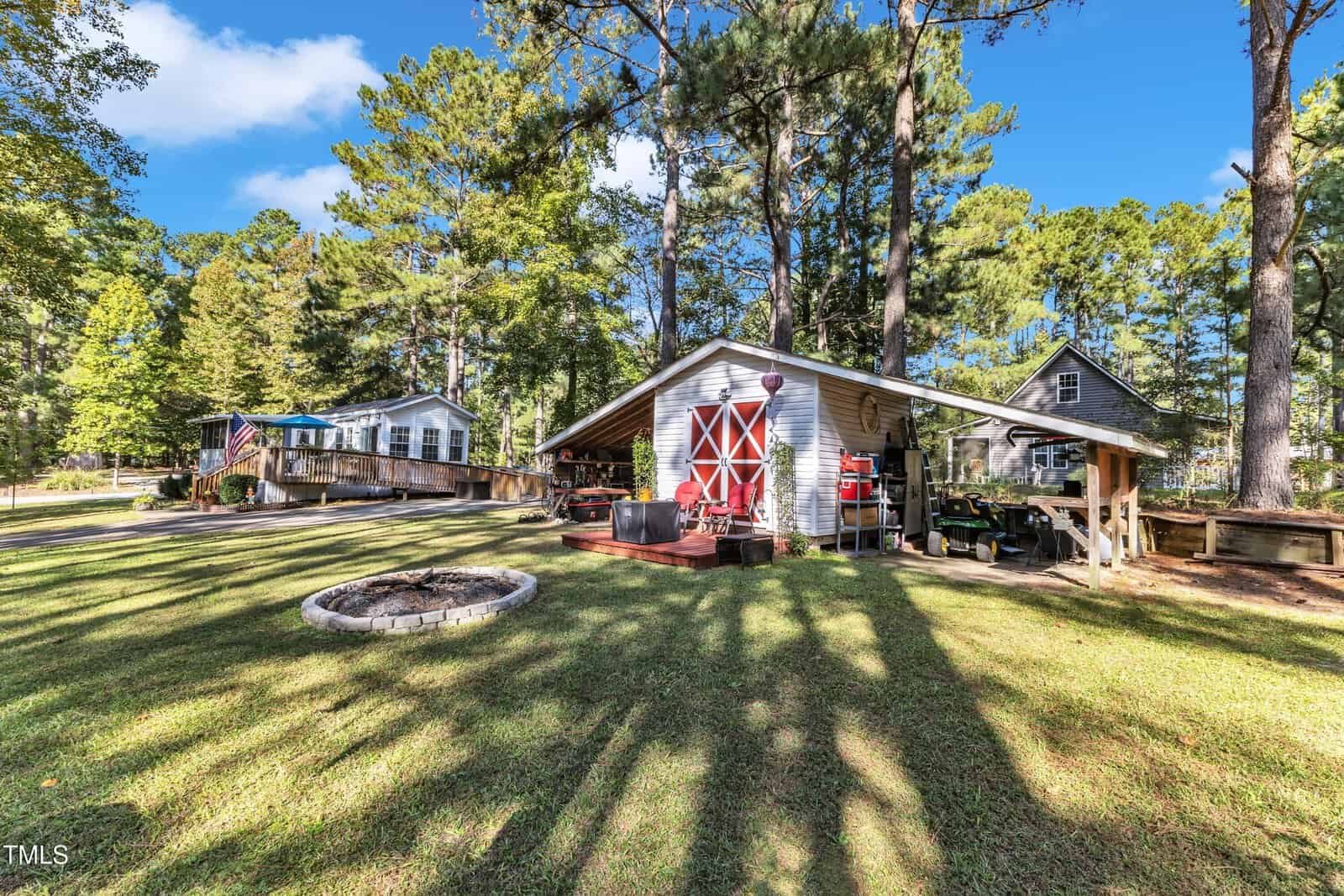 A yard with a fire pit, a large shed with a red double door, and two houses surrounded by tall trees under a sunny blue sky. Outdoor furniture and equipment are visible near the shed.