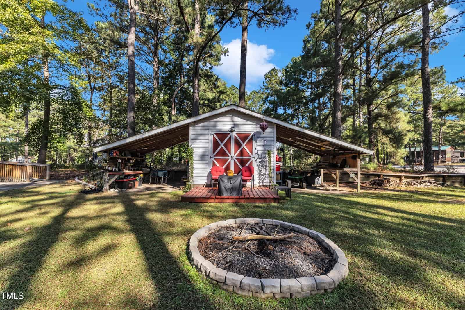 A small white shed with red double doors and a covered porch sits in a grassy yard surrounded by trees. There is a circular stone fire pit with charred wood in the foreground. Outdoor furniture is visible under the porch.