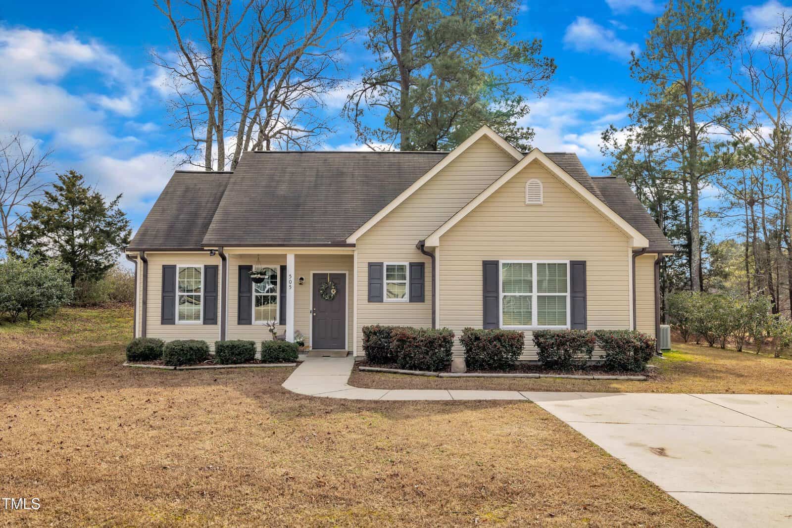 Located at 505 Wild Goose Lane in Creedmoor, this charming beige single-story house with dark shutters and a dark roof is surrounded by a dry lawn and sparse trees. A concrete driveway leads to the welcoming front entrance, adorned with a wreath on the door. The sky above is clear with a few clouds.