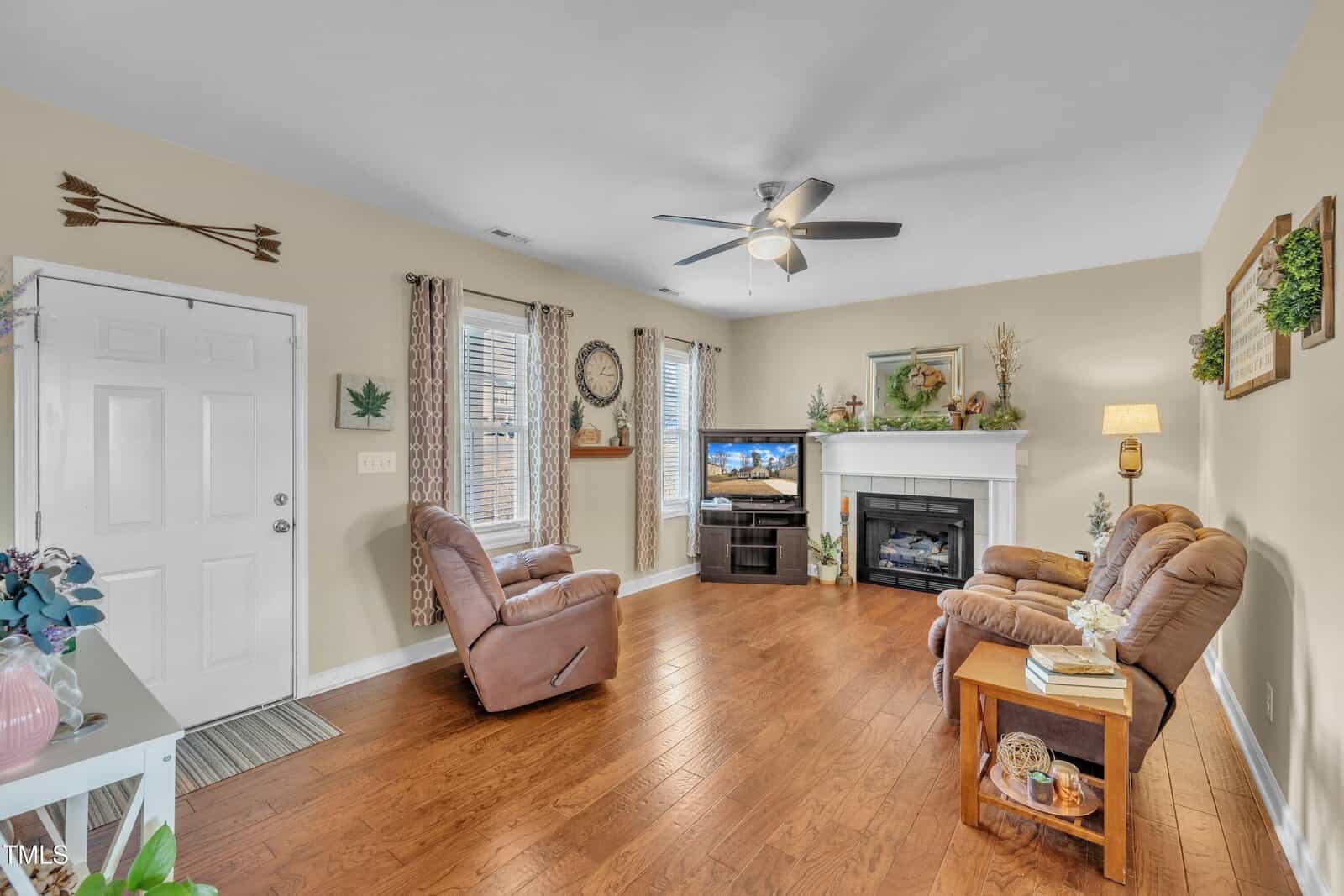 A cozy living room with hardwood floors, a ceiling fan, and light-colored walls. Two cushioned chairs face a fireplace with a TV above it. Decor includes plants, framed art, and wall clocks. Windows with curtains allow natural light in.