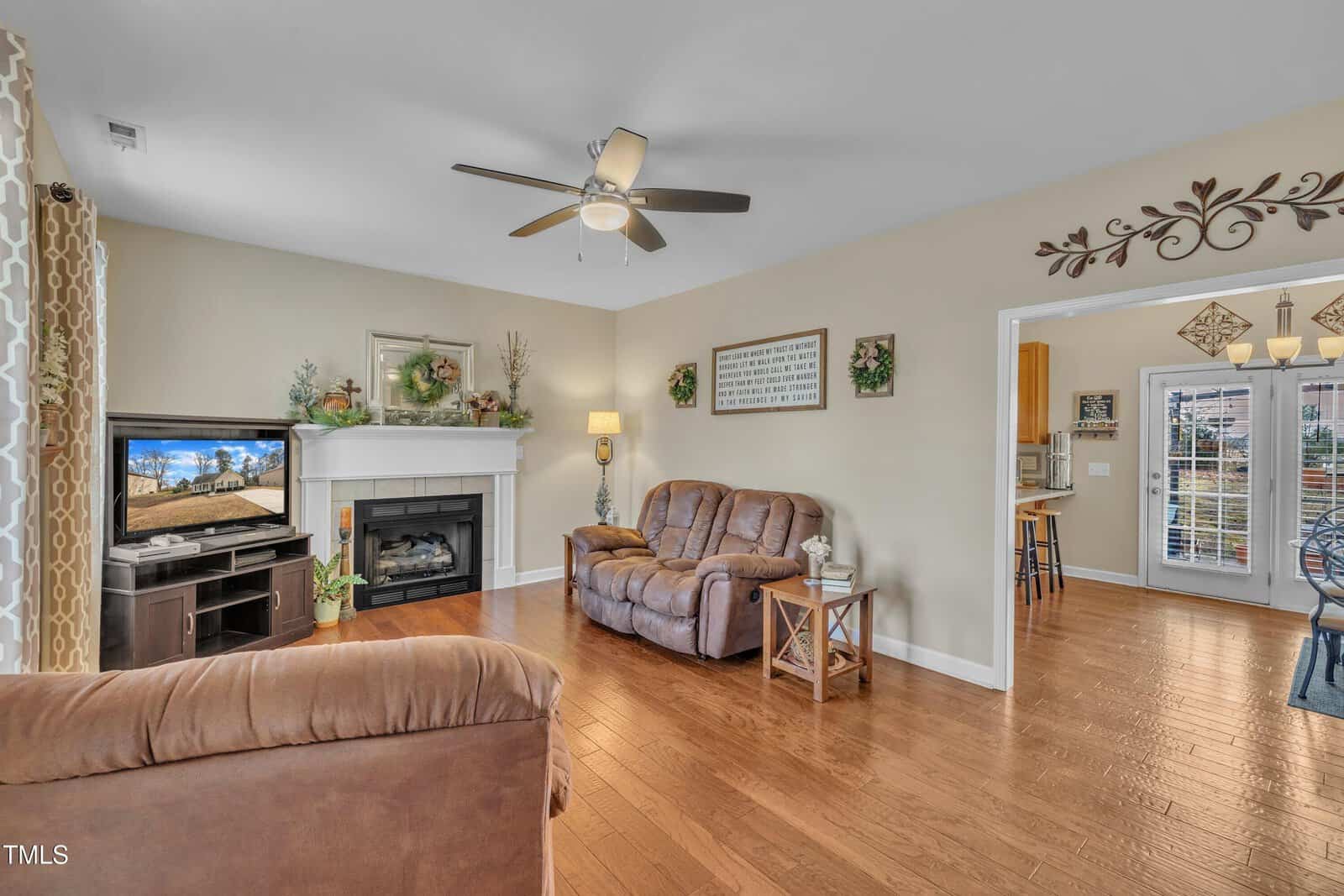 A living room with a brown sofa and recliner set, a TV on a stand next to a fireplace, and a ceiling fan. The room features wood flooring and decorative wall hangings. An open doorway leads to a kitchen area with a glass door to the outside.