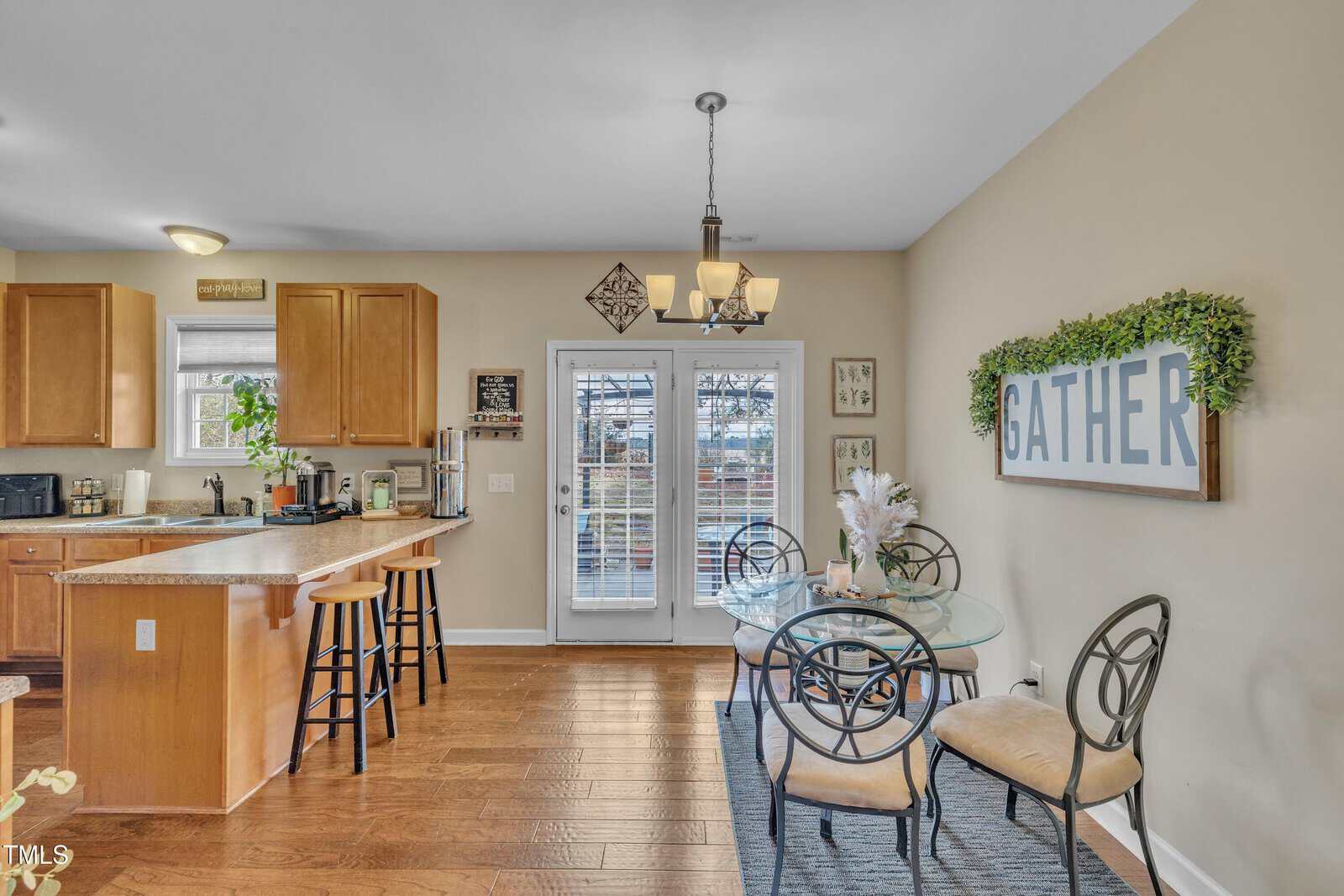Kitchen and dining area with wooden flooring. A kitchen island with stools, wooden cabinets, and appliances is on the left. A dining table with chairs is on the right. French doors lead outside, with a sign reading "GATHER" on the adjacent wall.
