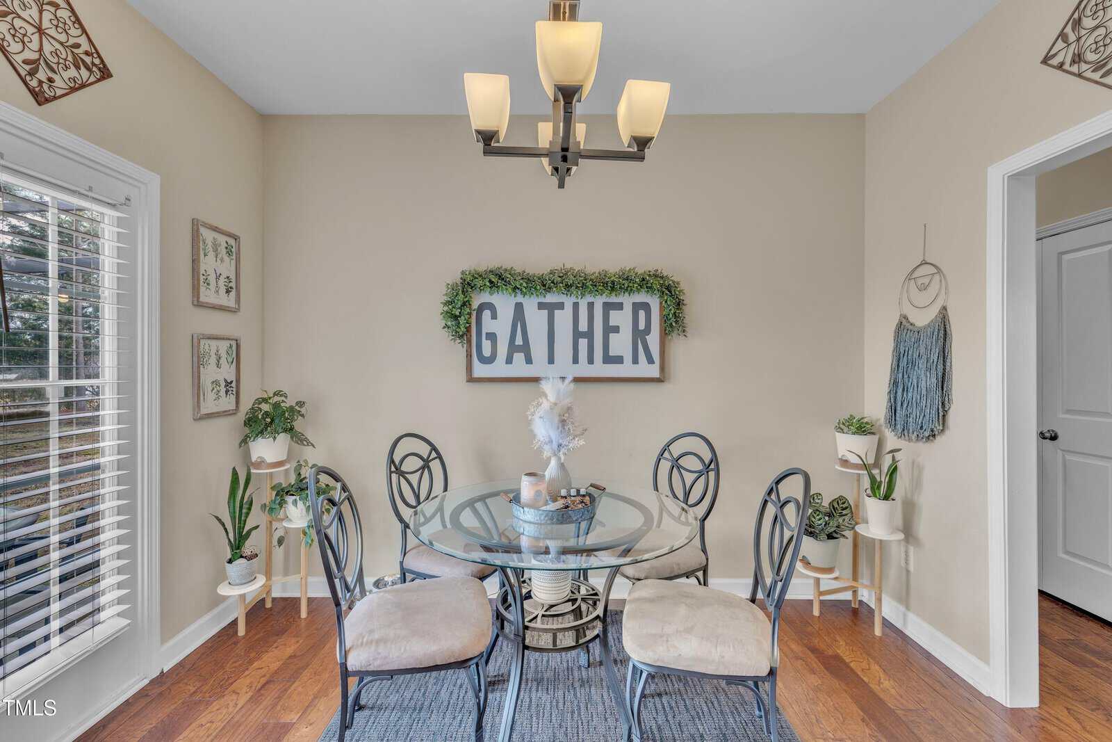 A dining area features a round glass table with four chairs on a blue rug. A sign reading "GATHER" is mounted on the wall, surrounded by greenery. A modern chandelier hangs above. Various plants and decorative items are placed around the room.