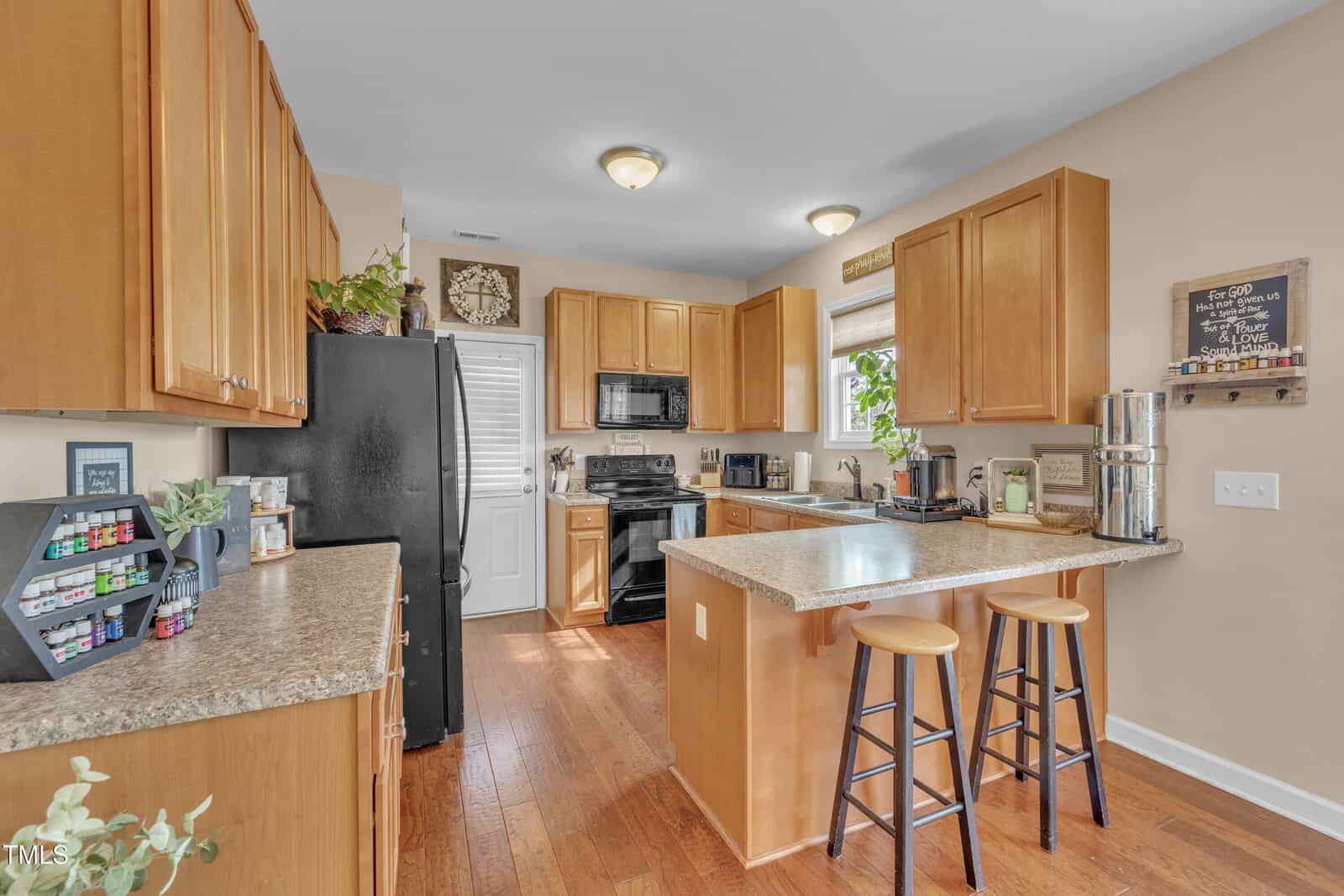 A modern kitchen with wooden cabinets and a black refrigerator. The countertop has two barstools, a toaster, a coffee maker, and various kitchen items. A plant and decorative items are on the cabinets. The floor is wood, and walls are beige.