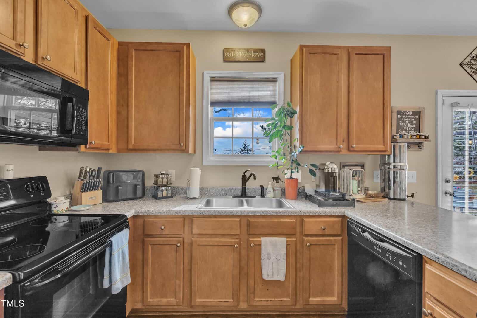 A kitchen with wooden cabinets and a granite countertop featuring a sink, houseplant, toaster, and coffee maker. A stove and microwave are on the left, and a small window with a view outside is above the sink.