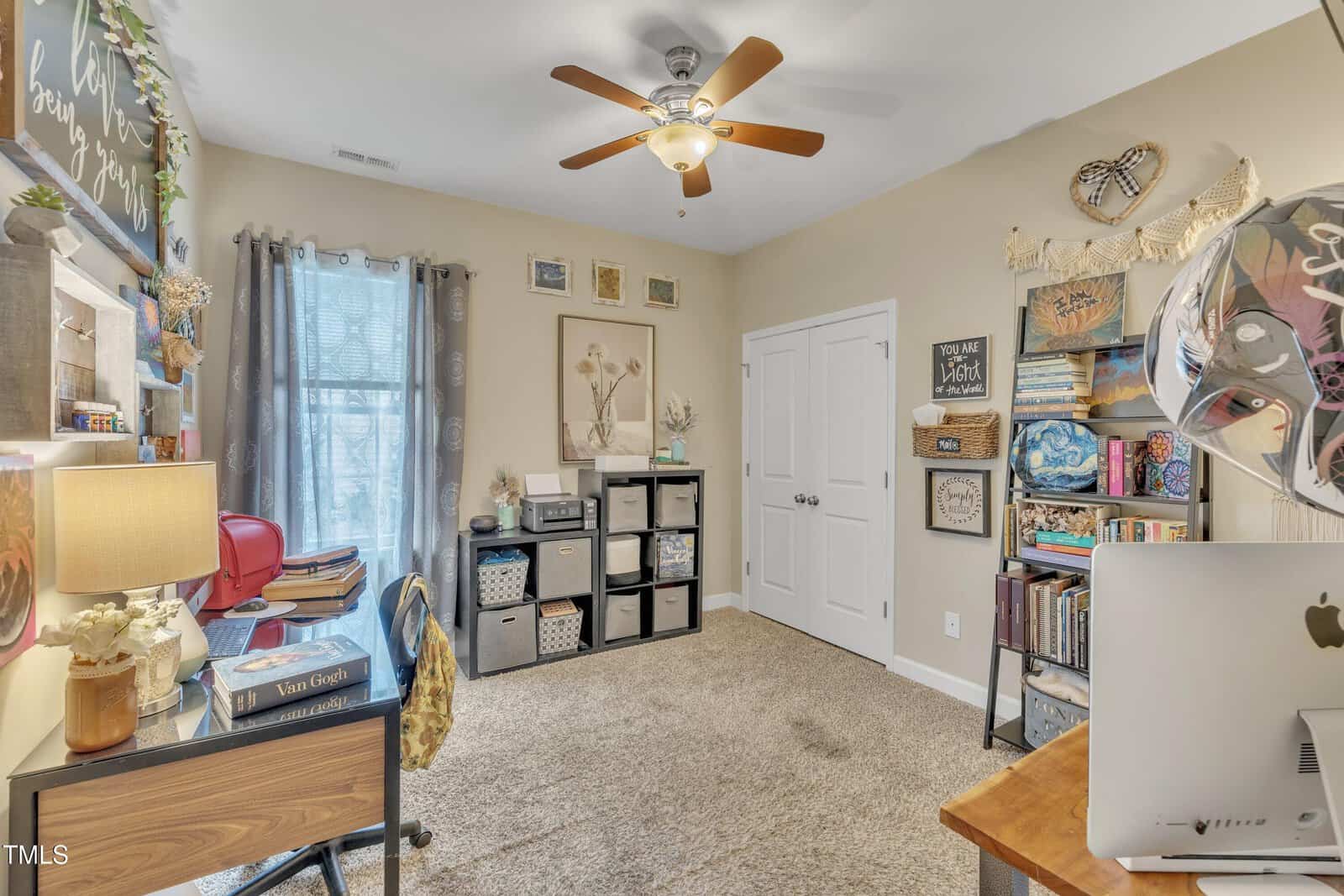 A home office with beige walls, a ceiling fan, and carpeted floor. It features a desk with a computer, a chair, bookshelves filled with books and decor, a window with curtains, and wall art. A potted plant sits on the desk.