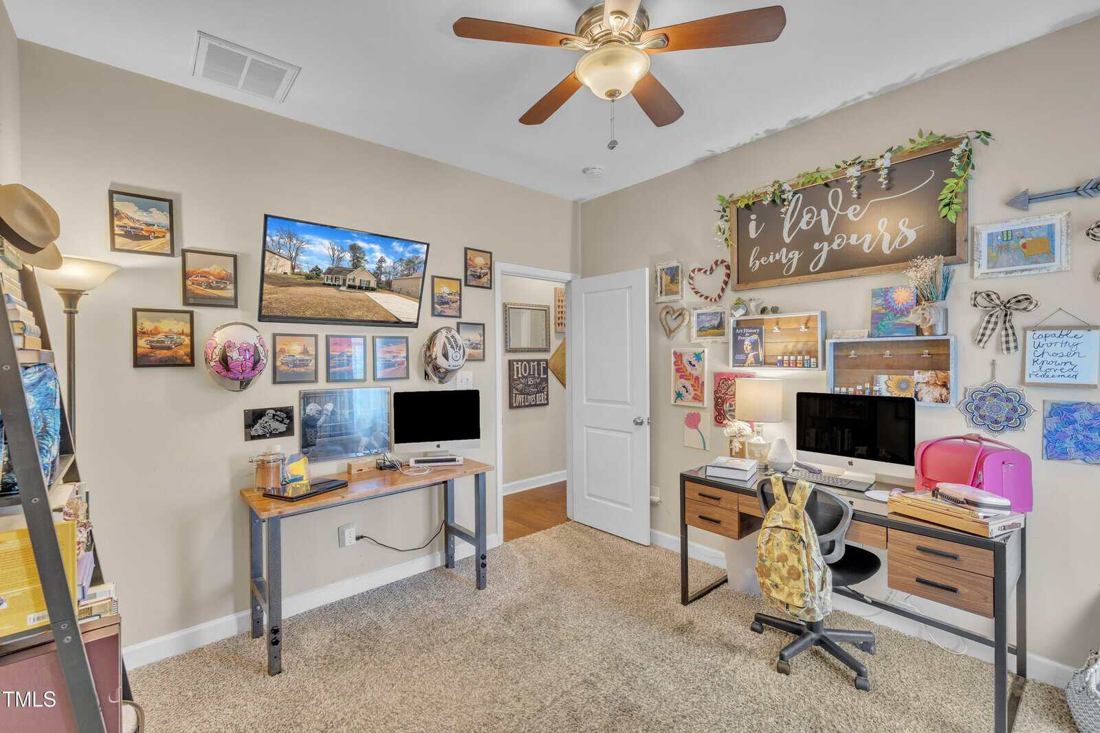 A home office with two wooden desks, each holding a computer. Walls are adorned with various pictures and decor, including a large sign reading "love bring yours." A ceiling fan is above, and a closed white door is in the background. Carpeting covers the floor.
