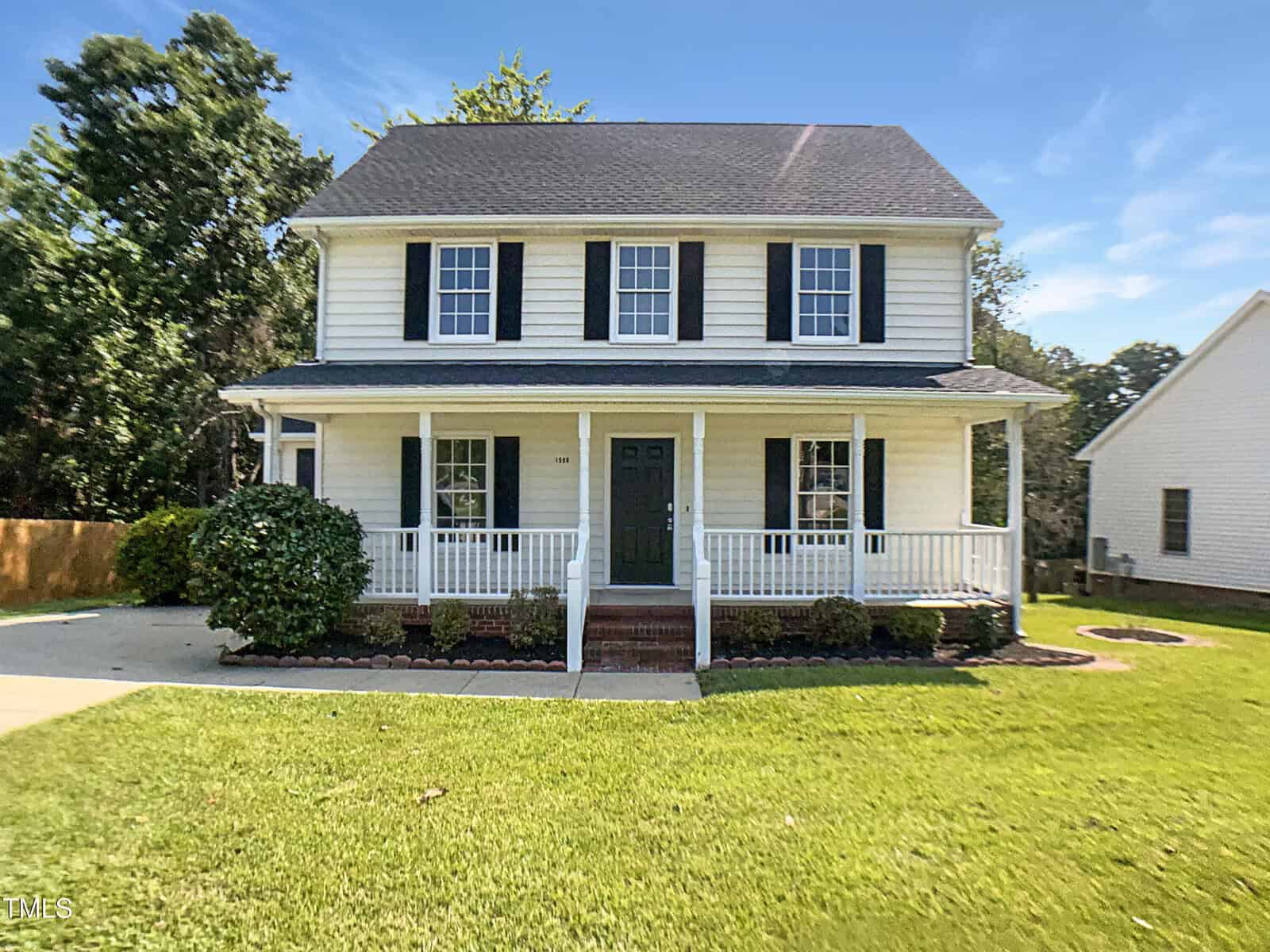 A two-story white house with black shutters and a covered front porch at 1599 Rogers Pointe Lane, Creedmoor, sits on a well-manicured lawn with shrubs and a paved driveway. Trees are visible in the background under a blue sky.