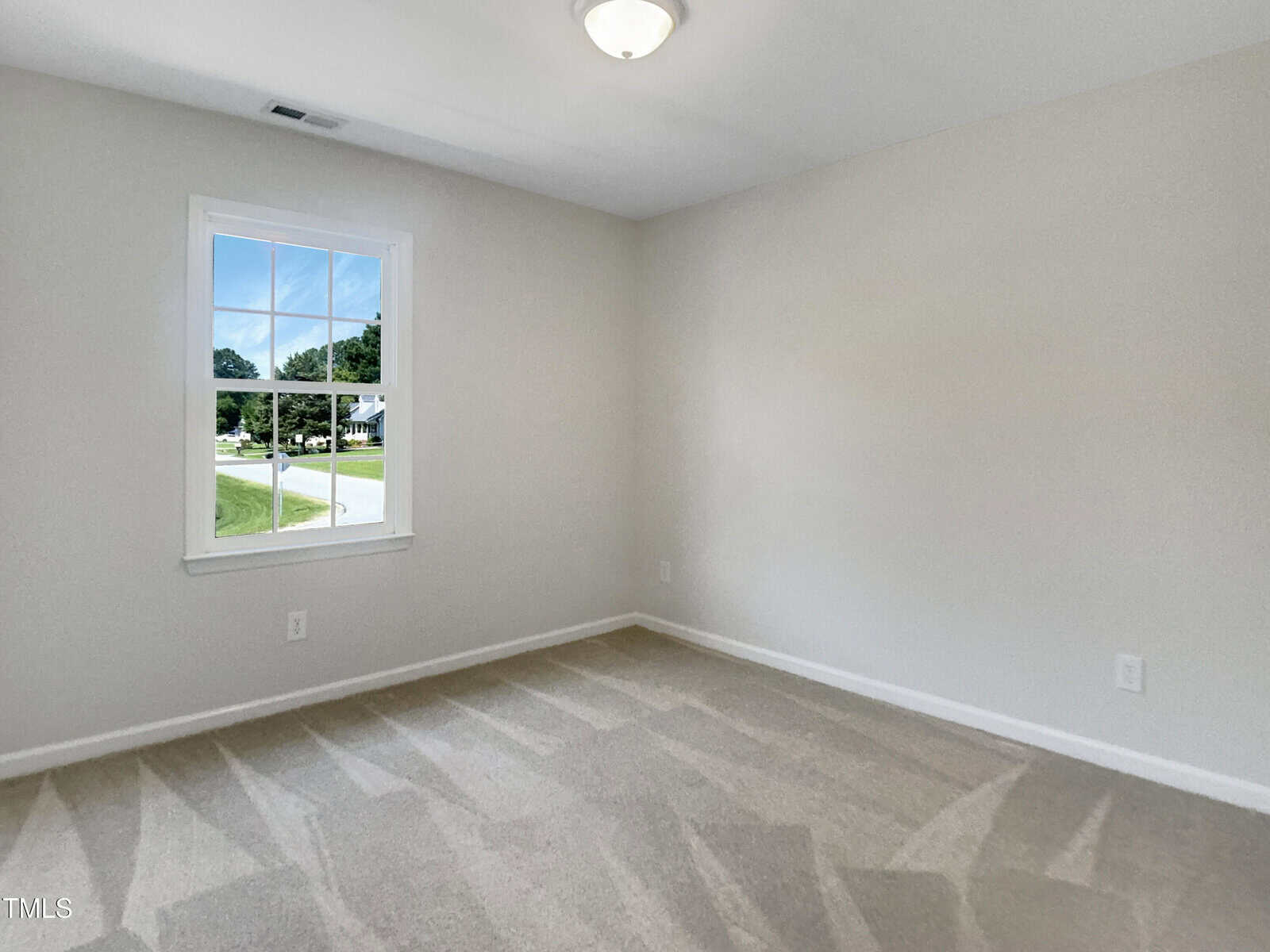 An empty room with beige carpet, light gray walls, a single window showing a street and green trees outside, and a ceiling light fixture. The space appears clean and well-lit.