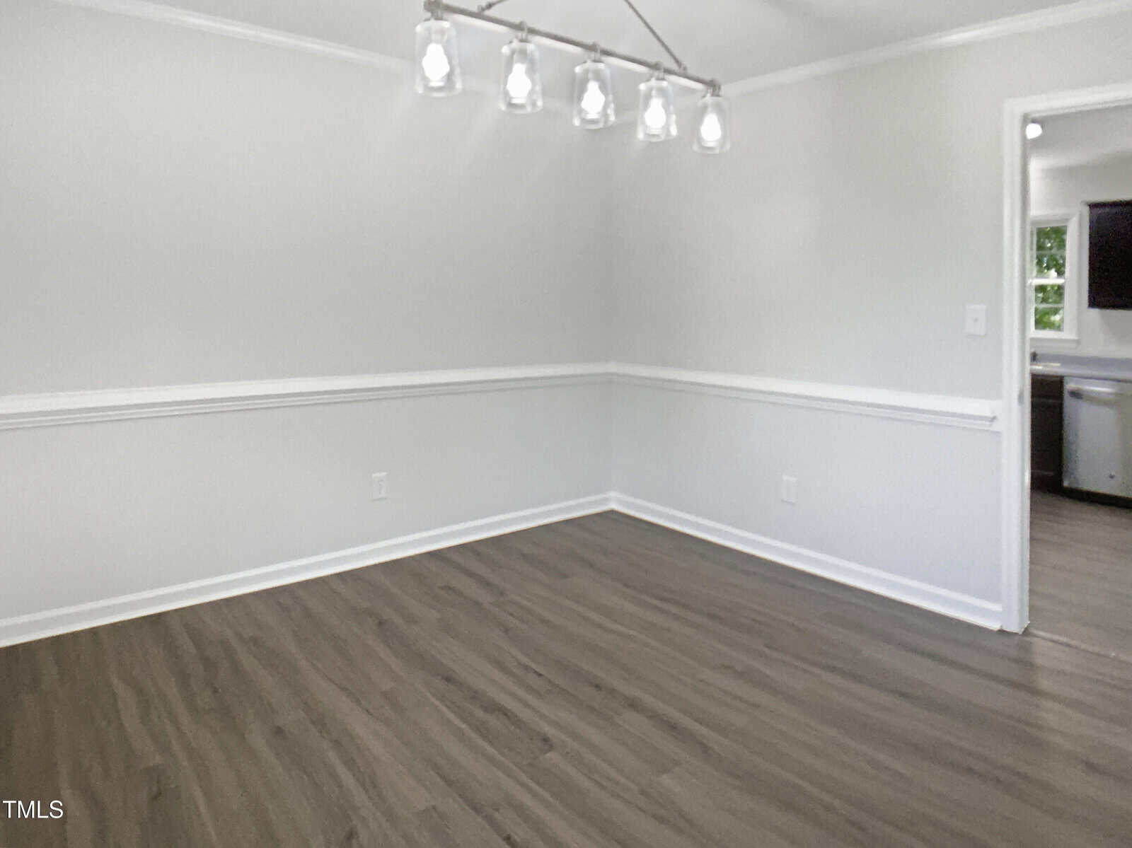 An empty room with light gray walls, white trim, wood-look flooring, and a modern ceiling light fixture. An open doorway leads to a kitchen with visible stainless steel appliances.