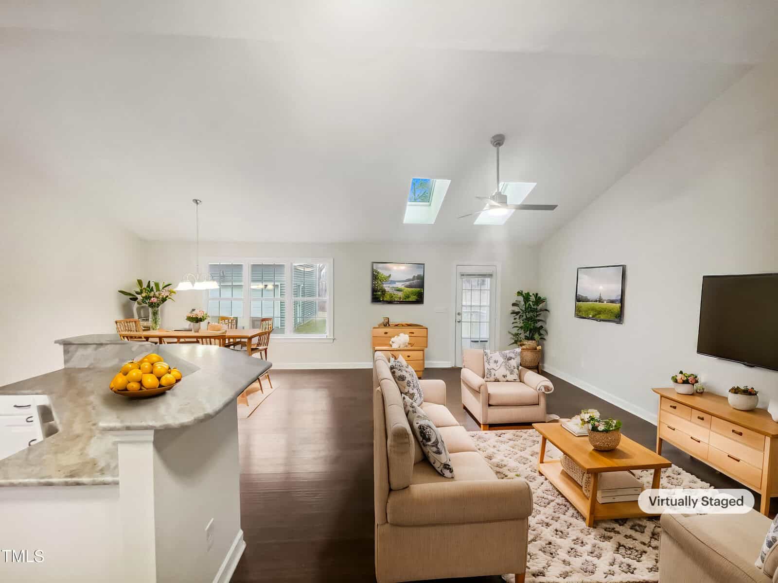 Virtually staged image of a bright, open living space. It features a dining area, stone countertop with a bowl of oranges, beige sofas, a TV, and skylights. Decorative plants and framed pictures are placed throughout the room.