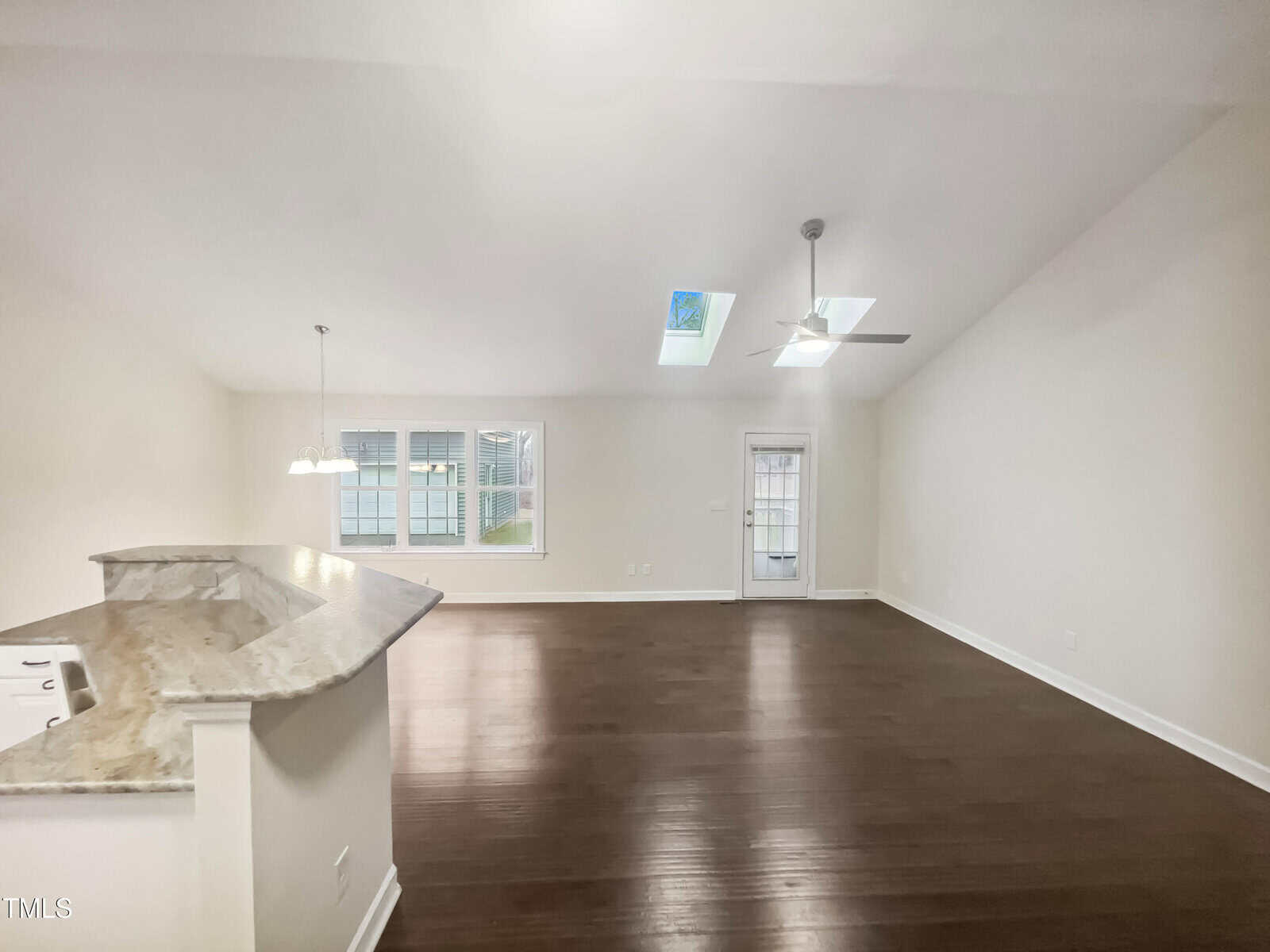 A spacious, empty room with dark wood floors and white walls. The room features a vaulted ceiling with skylights, a ceiling fan, and a small chandelier. There's a curved countertop on the left and a door leading outside in the background.