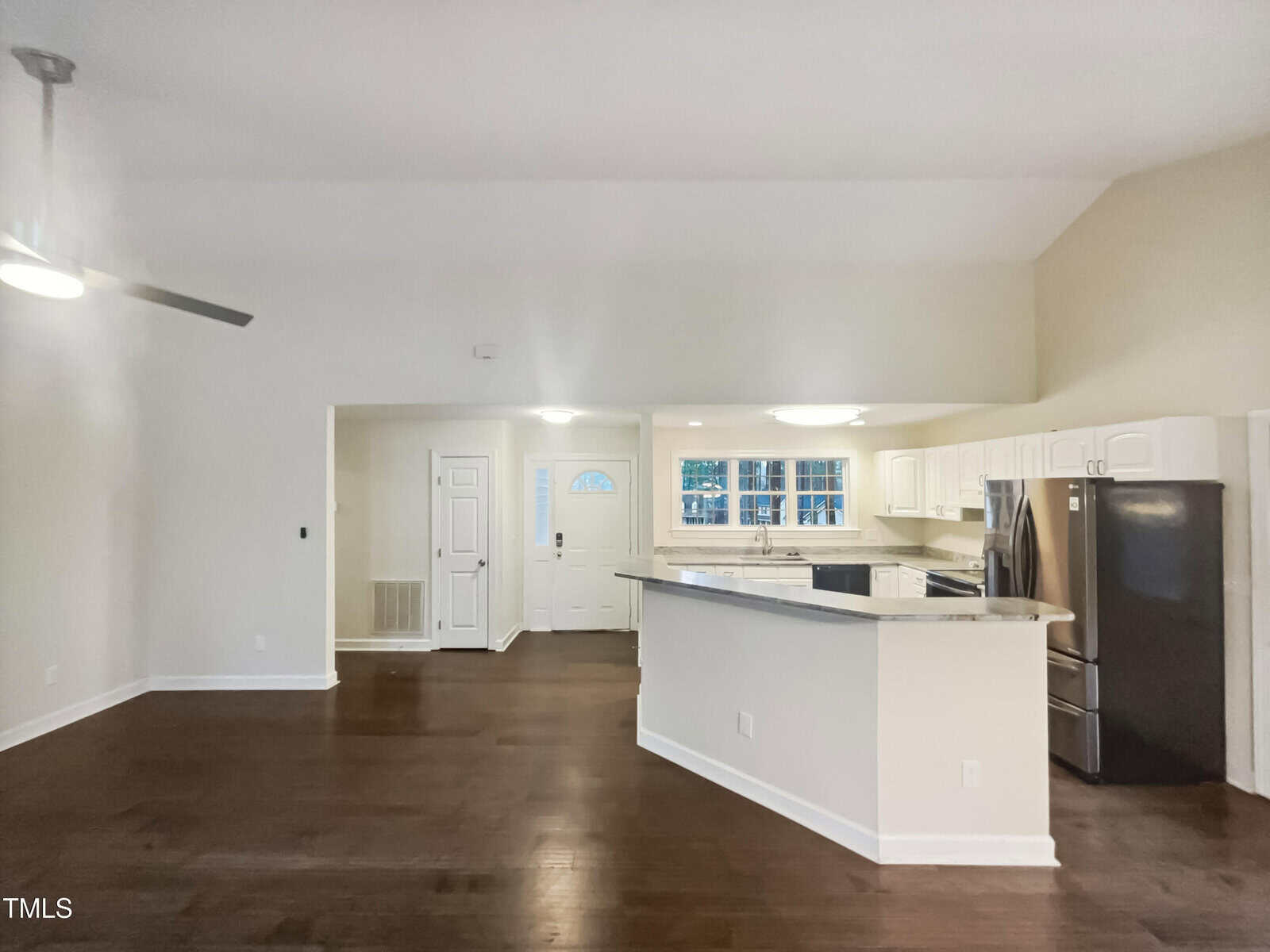 Interior view of an unfurnished room featuring hardwood floors and white walls. A partially visible kitchen with a countertop, stainless steel appliances, and a row of windows is in the background. A ceiling fan is on the left side of the room.