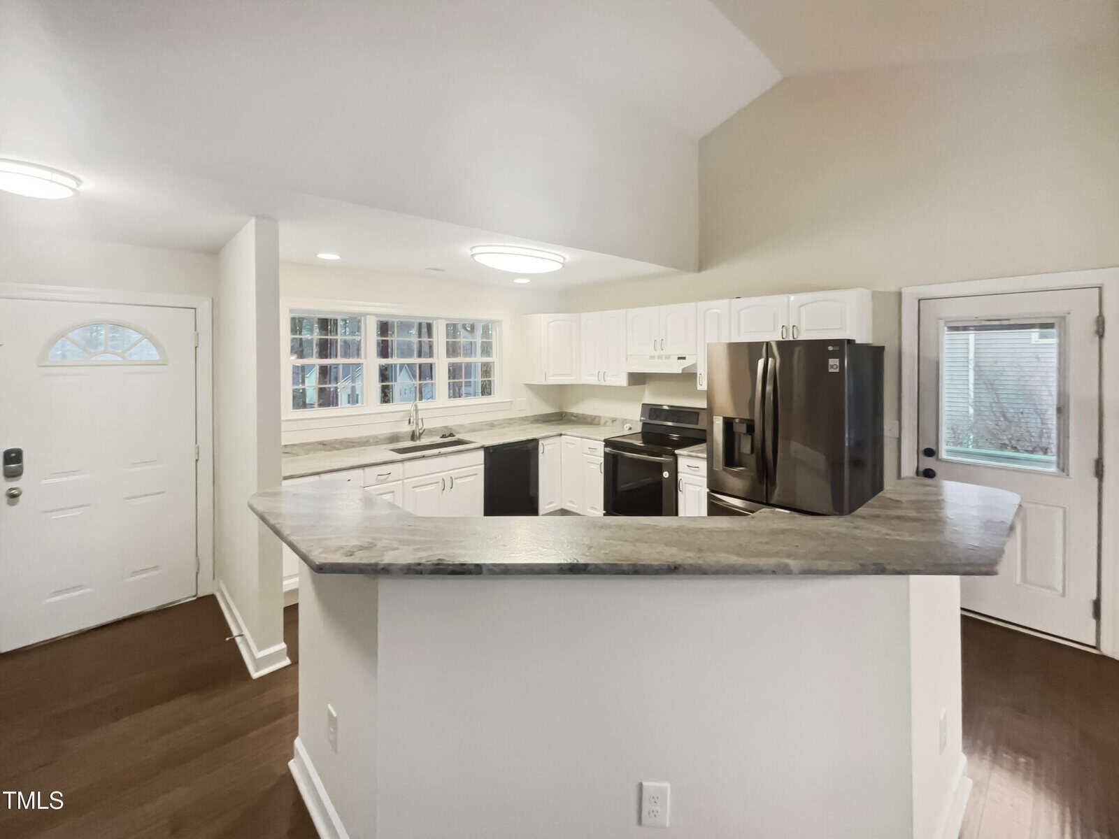 Modern kitchen with white cabinets, marble countertops, and stainless steel appliances, including a refrigerator and oven. A curved counter extends into the room, and there is a door and multiple windows providing natural light.