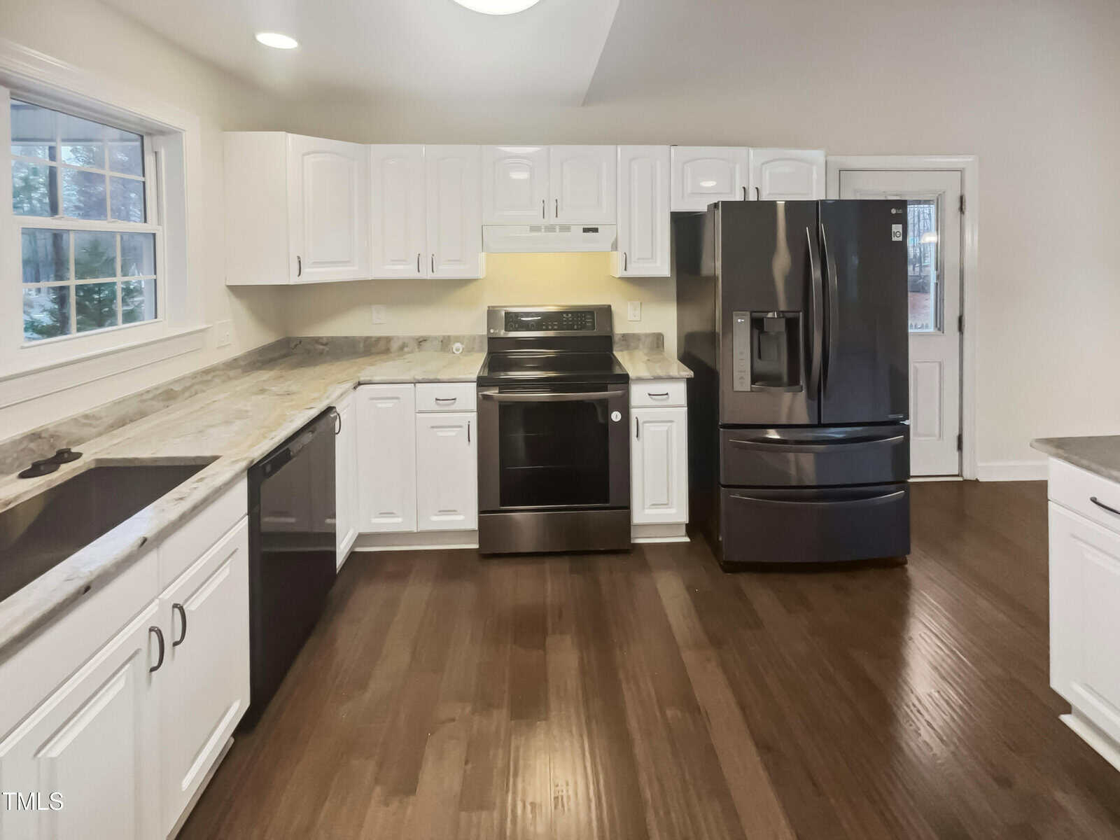 A modern kitchen featuring white cabinets, a black refrigerator, and a black stove with an overhead vent. The countertops are a light-colored granite, and the floor is wooden. There is a window above the sink and a door leading outside.