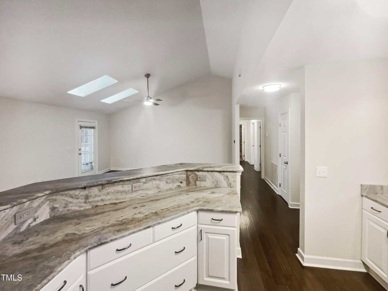 A kitchen with marble countertops and white cabinets. The room features a curved counter, wood flooring, white walls, and a ceiling fan. Skylights are visible above, and several doorways lead to adjacent rooms.