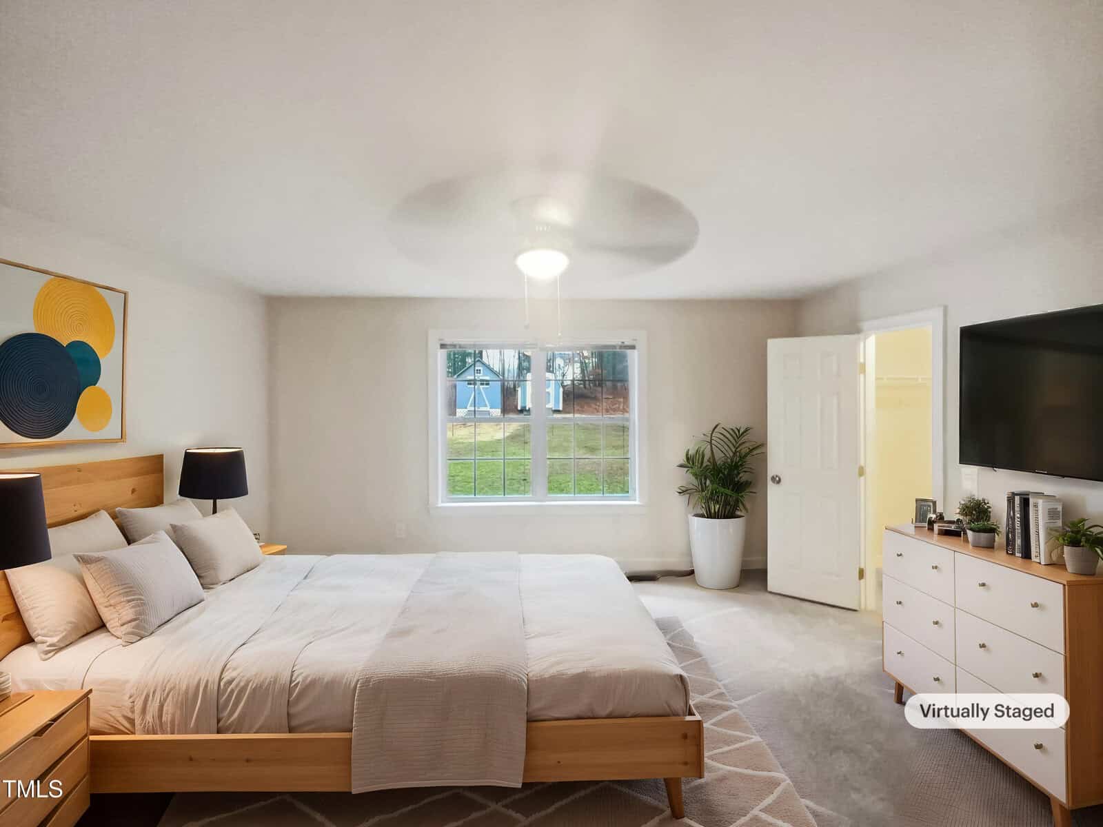 A bedroom with a large bed featuring white and beige bedding, two black lamps on bedside tables, a dresser with a plant and decorative items, and a window overlooking a yard. The walls are light-colored, and there is a ceiling light fixture.