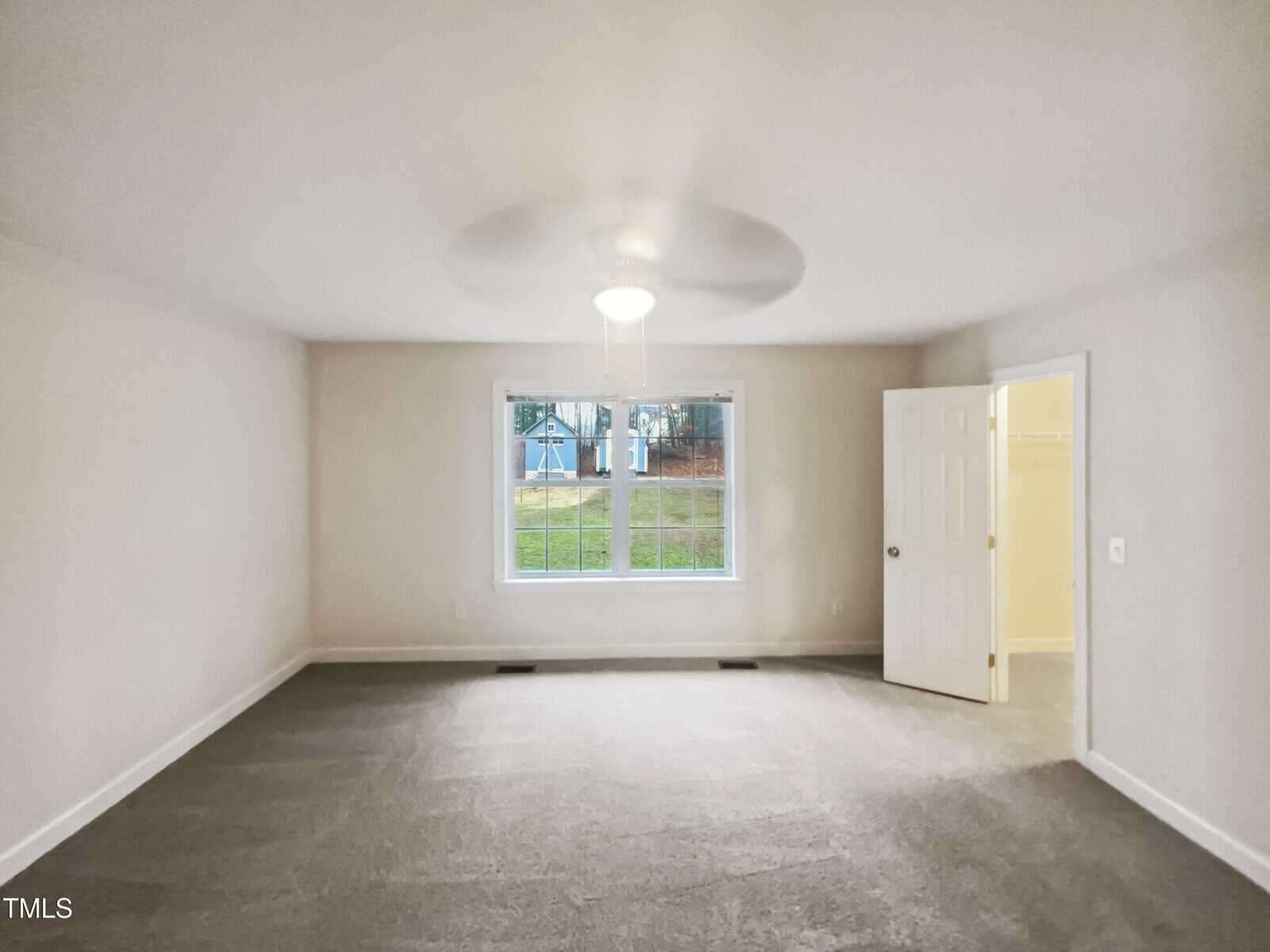 An empty room with light gray carpet and off-white walls features a large window facing outside. A ceiling fan with a light is mounted in the center, and an open door leads to a closet. Natural light illuminates the space.